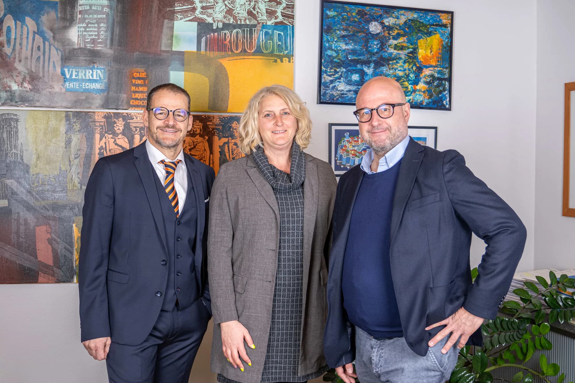 Drei Mitglieder der Kanzlei stehen nebeneinander im Büro vor einer Wand mit farbigen gerahmten Bildern und blicken in die Kamera.
