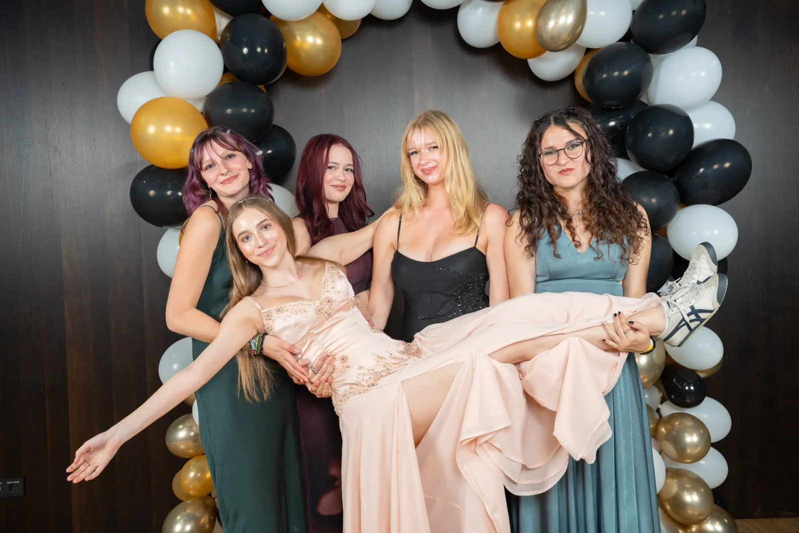 Vier Freundinnen tragen eine Freundin im rosa Kleid quer vor dem Ballonbogen