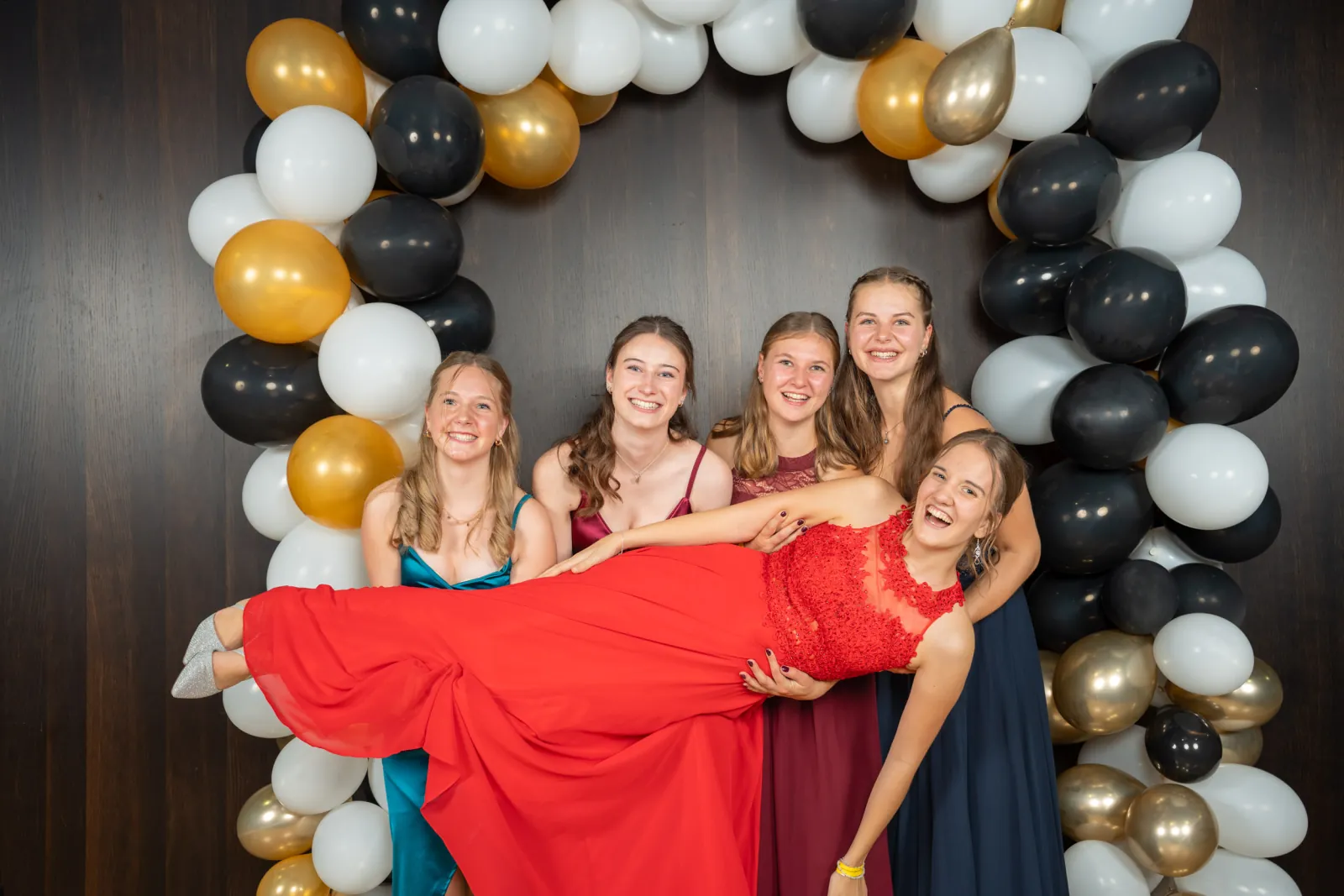 Freundinnen halten eine junge Frau im roten Kleid quer vor dem Ballonbogen