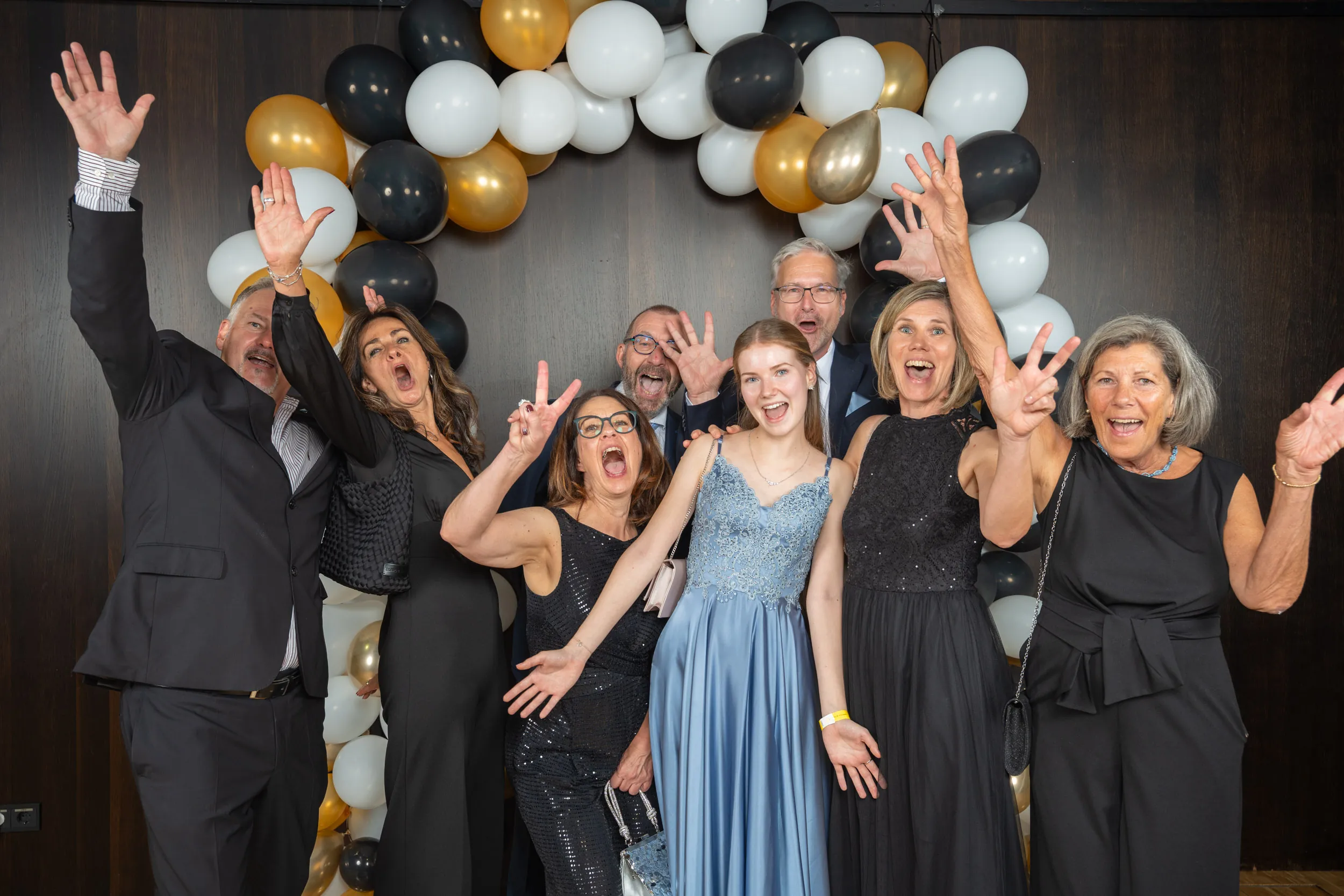 Ballgäste reißen vor dem schwarz-weiß-goldenen Ballonbogen der Fotostation lachend die Arme hoch