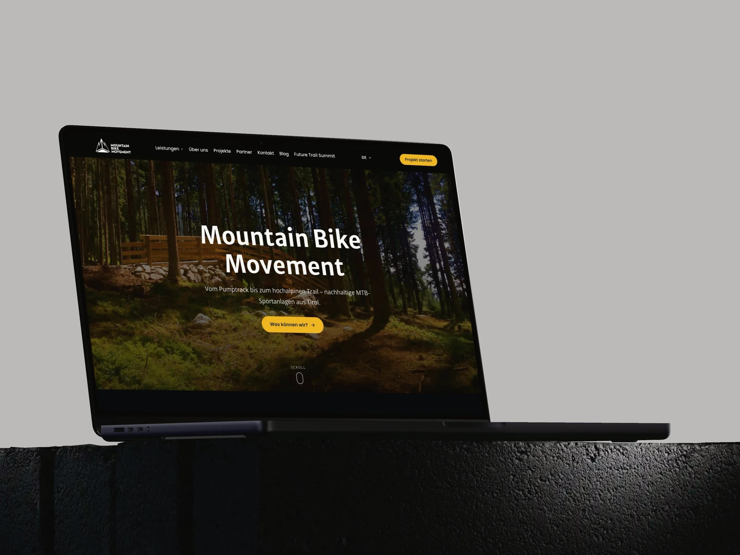Ein MacBook Pro auf einem dunklen Steinsockel vor hellgrauem Grund zeigt die Startseite von mountainbikemovement.com mit einem Waldtrail und dem Schriftzug Mountain Bike Movement.