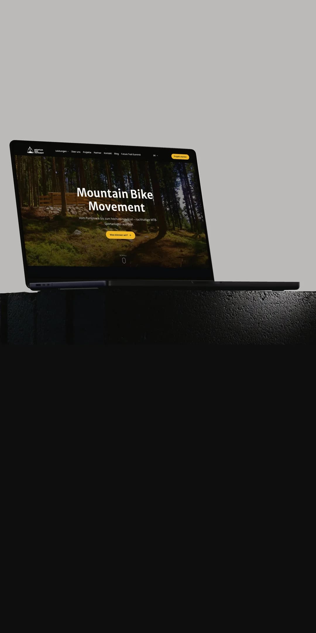 Ein MacBook Pro auf einem dunklen Steinsockel vor hellgrauem Grund zeigt die Startseite von mountainbikemovement.com mit einem Waldtrail und dem Schriftzug Mountain Bike Movement.