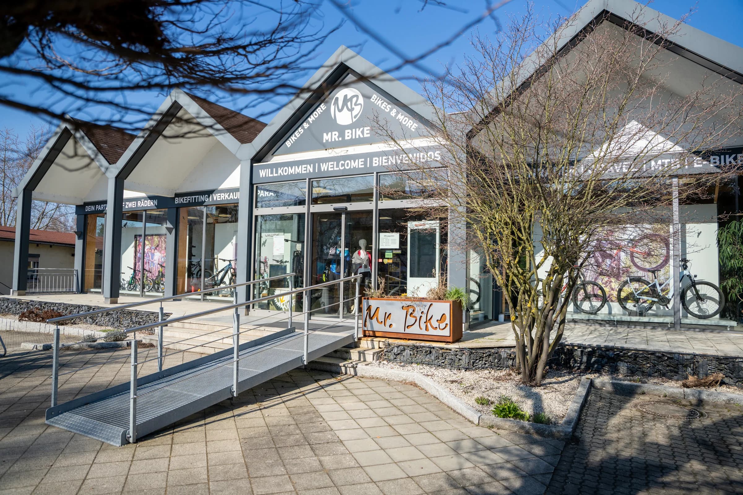 Der Eingang des Mr. Bike Stores schräg von vorn: die Rampe mit Geländer, das Signet über der Tür, ein Baum und der gepflasterte Vorplatz in der Sonne.