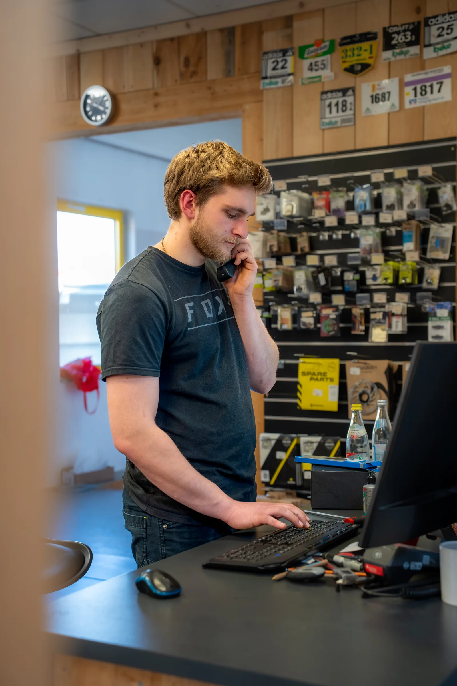 Ein Mechaniker im schwarzen T-Shirt telefoniert am Tresen der Werkstatt, hinter ihm eine Wand mit Ersatzteilen und Startnummern.