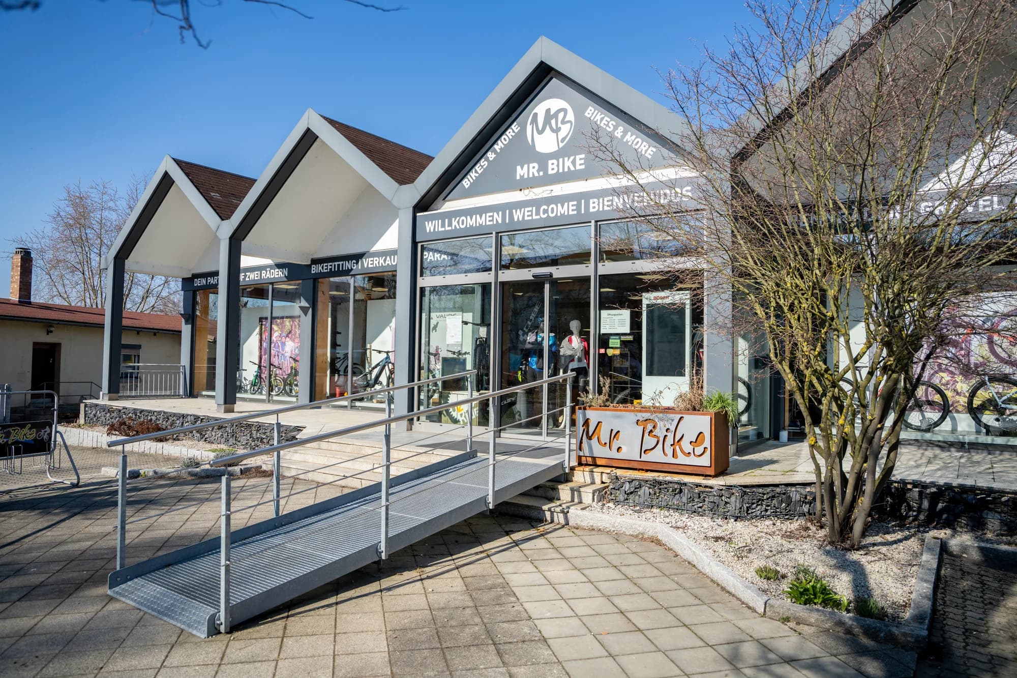 Der Eingang des Mr. Bike Stores mit Rampe und Geländer, dem MB-Signet über der Tür und einem Baum davor, im Schaufenster ein Rad.