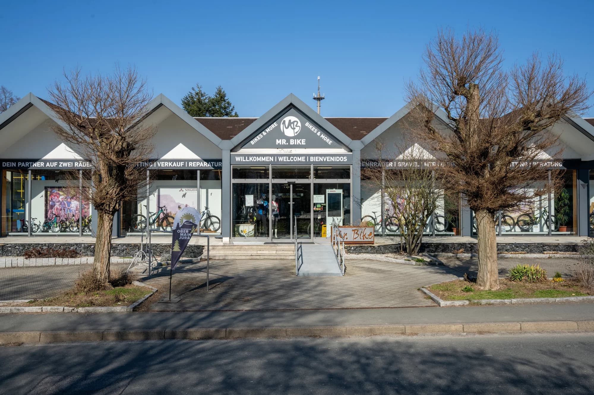 Die Front des Mr. Bike Stores in Lichtenfels mit den Spitzgiebeln und dem Schriftzug WILLKOMMEN WELCOME BIENVENIDOS, davor zwei kahle Bäume und blauer Himmel.