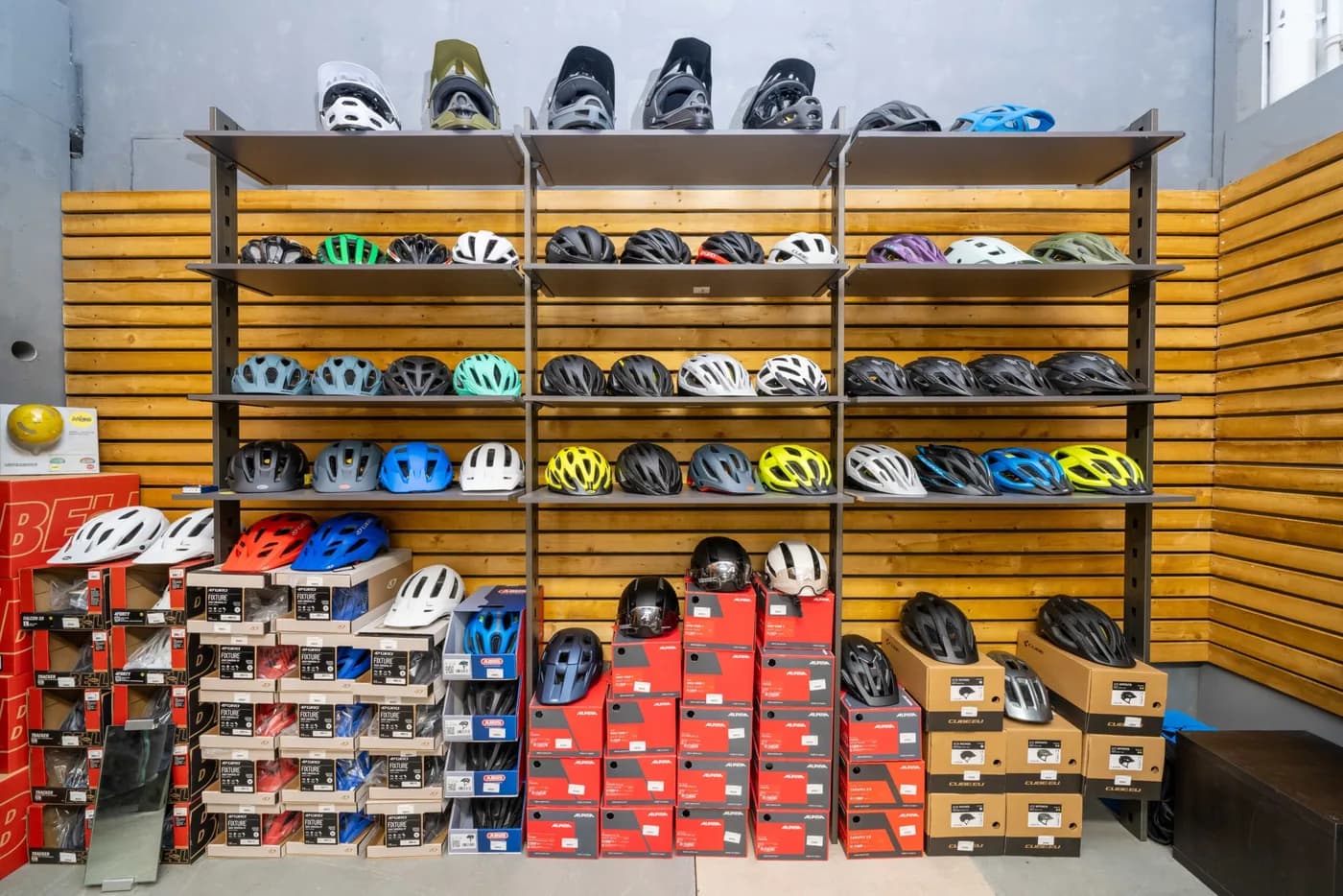 Eine Wand aus Holzfächern mit Fahrradhelmen in vielen Farben, darunter gestapelte Schuhkartons.