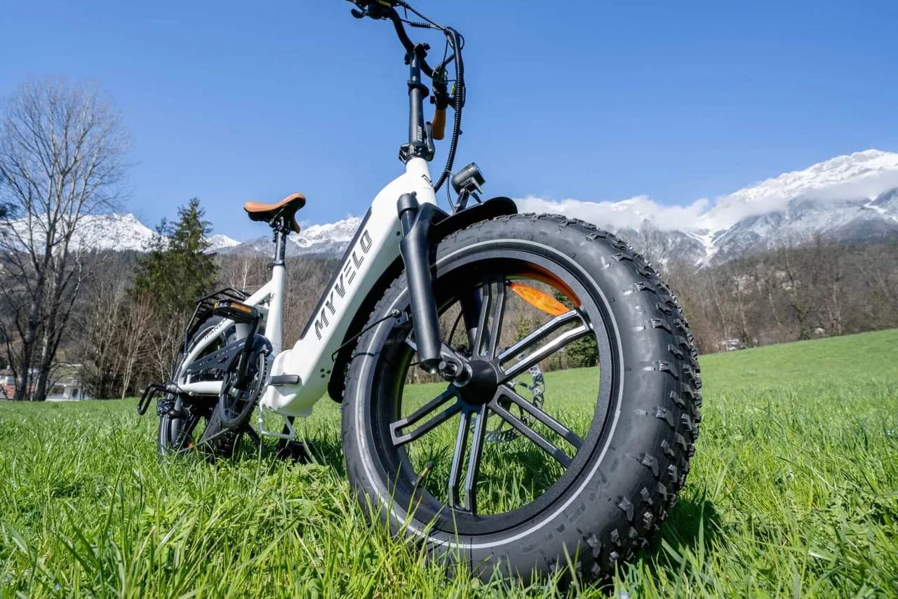 Ein weißes E-Klapprad steht im Gras, dicht am Vorderrad aufgenommen, dahinter die verschneiten Berge über Innsbruck.