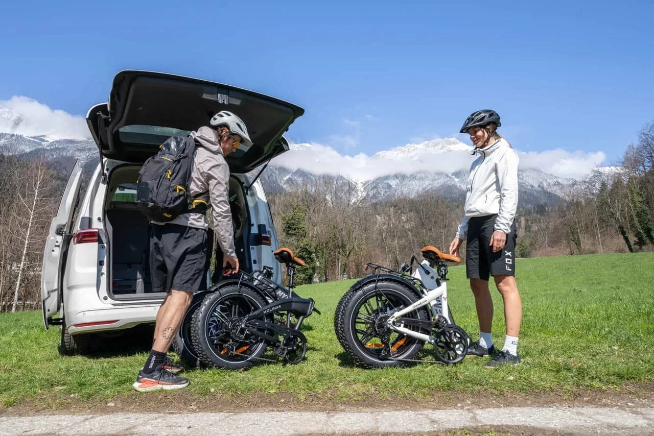 Zwei Personen laden gefaltete E-Klappräder in den Kofferraum eines weißen Vans, dahinter eine Wiese und verschneite Berge.