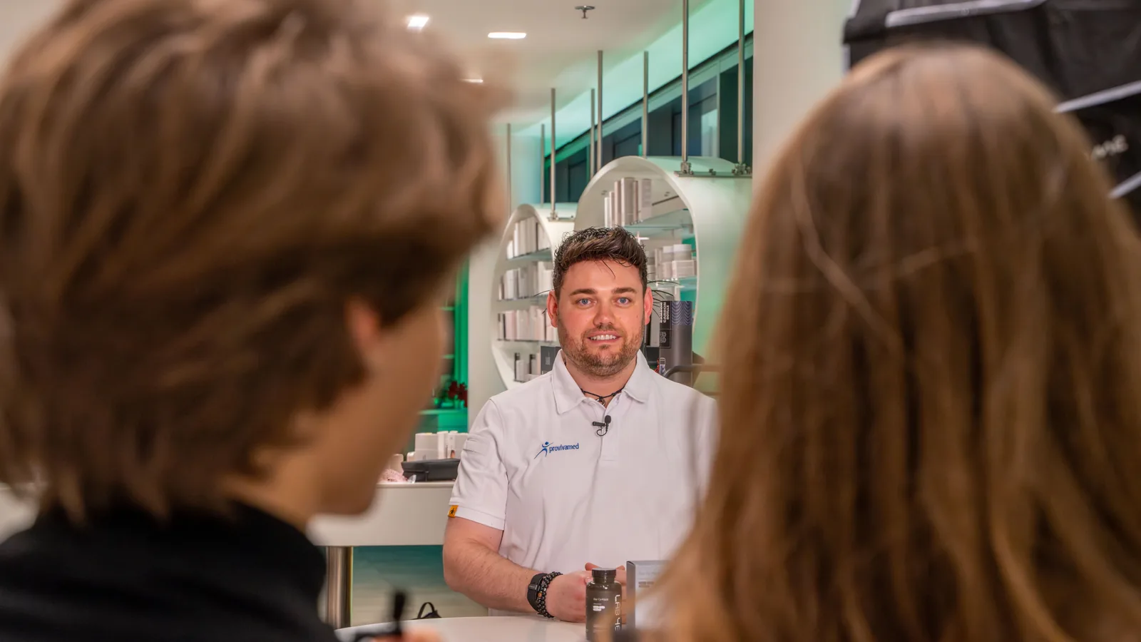 Beratung am Tresen einer Apotheke, über die Schulter der Kundschaft gesehen: dahinter ein Mitarbeiter im provivamed-Polo an der runden Theke.