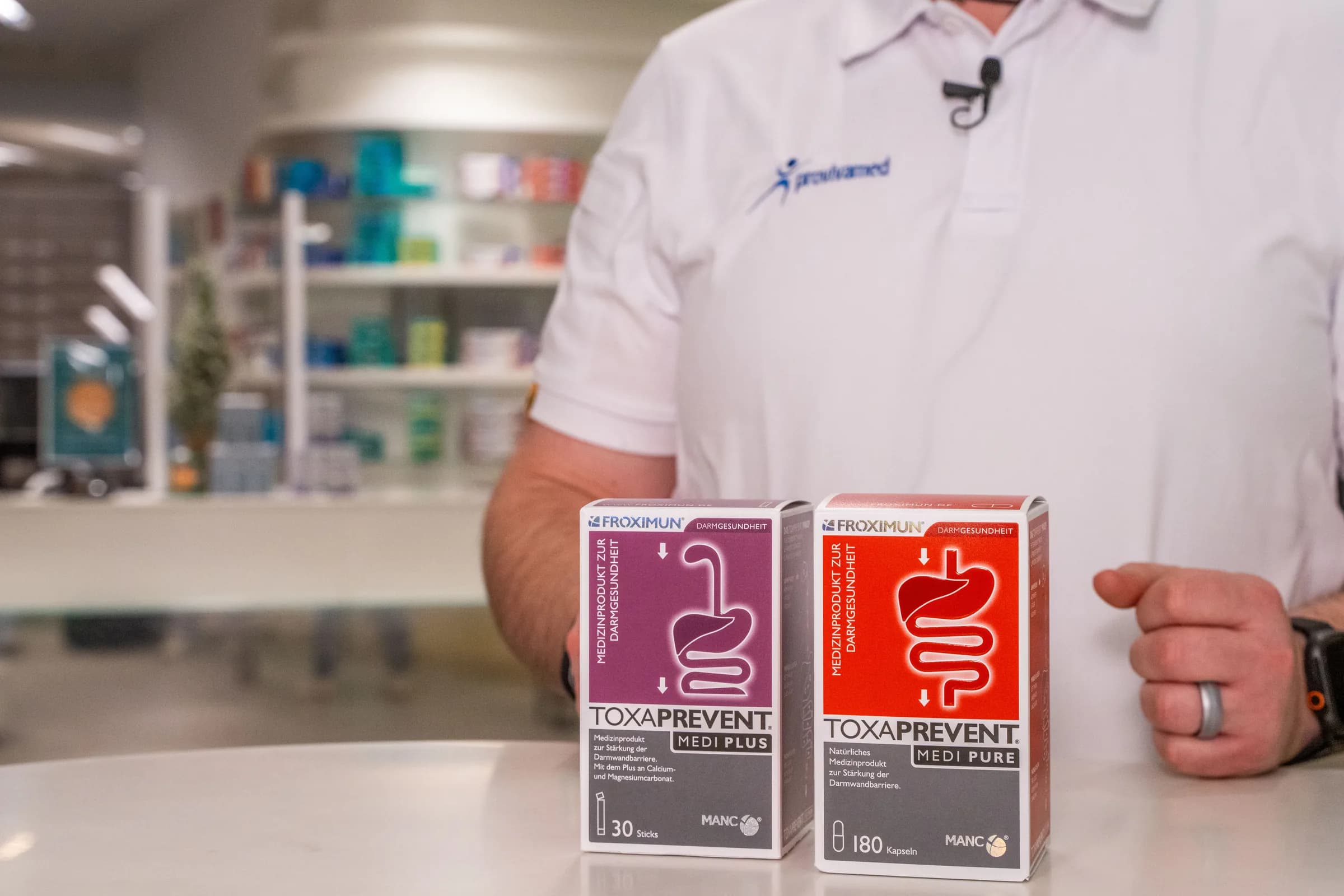 Ein Mitarbeiter hält zwei Froximun-Packungen in die Kamera, dahinter die Regalwand der Apotheke.