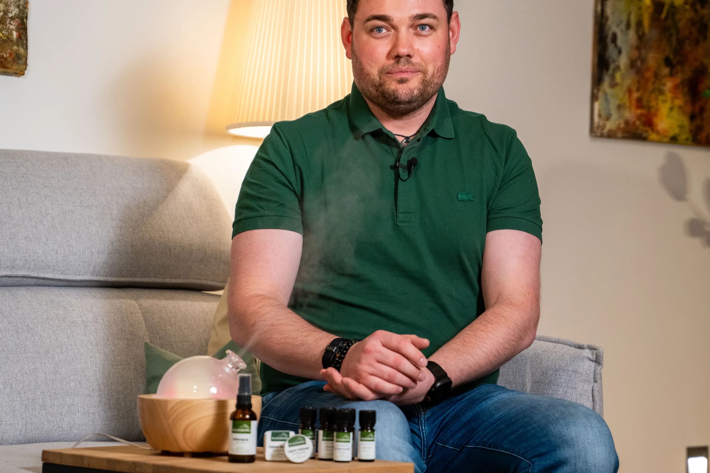 Ein Mann im grünen Polo sitzt auf einem Sofa, vor ihm auf dem Beistelltisch der Diffuser und die Öle von KLEEBERG.