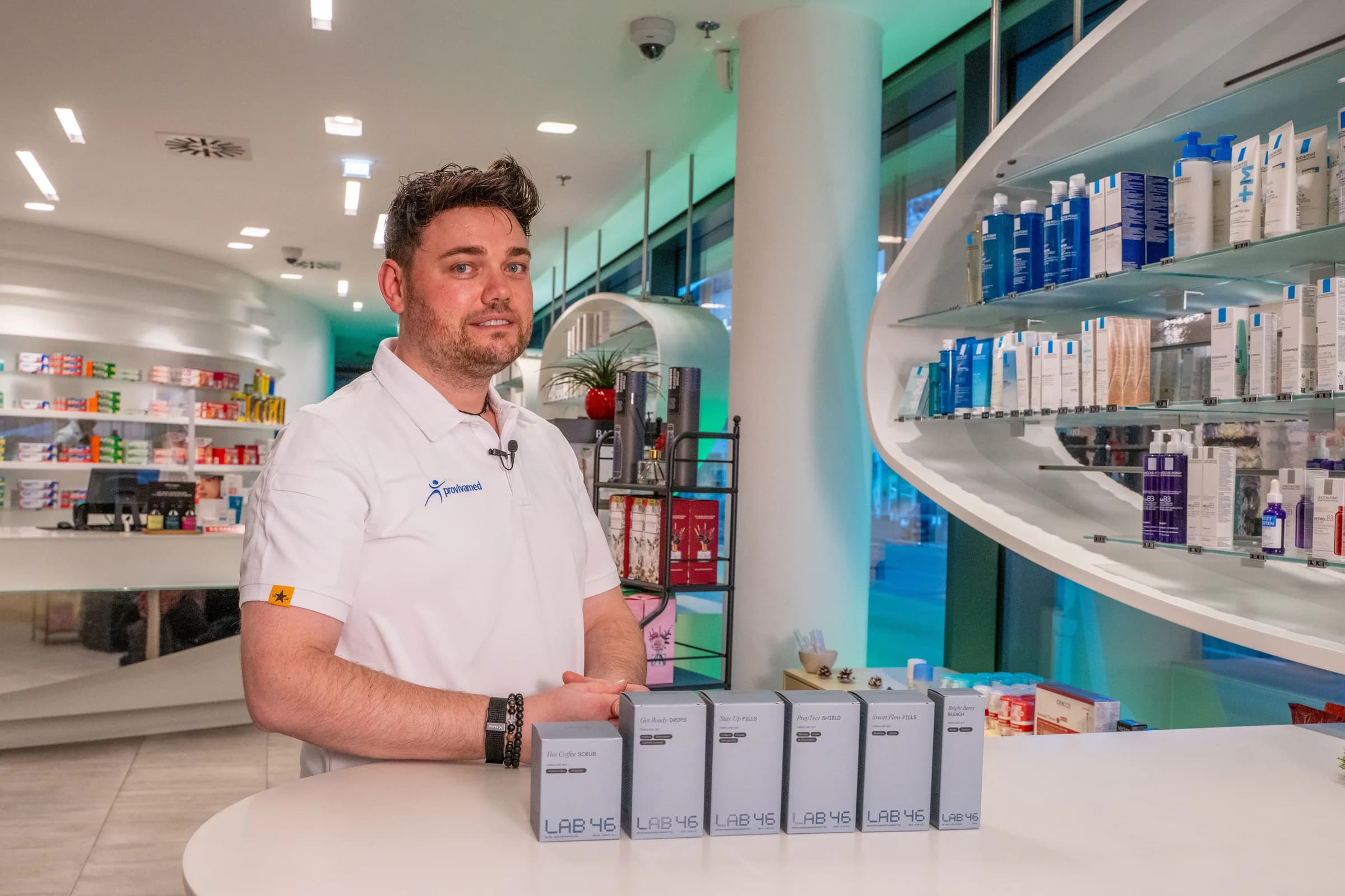 Ein Mitarbeiter im weissen provivamed-Polo steht an der runden Theke einer Apotheke, davor eine Reihe LAB46-Kartons.