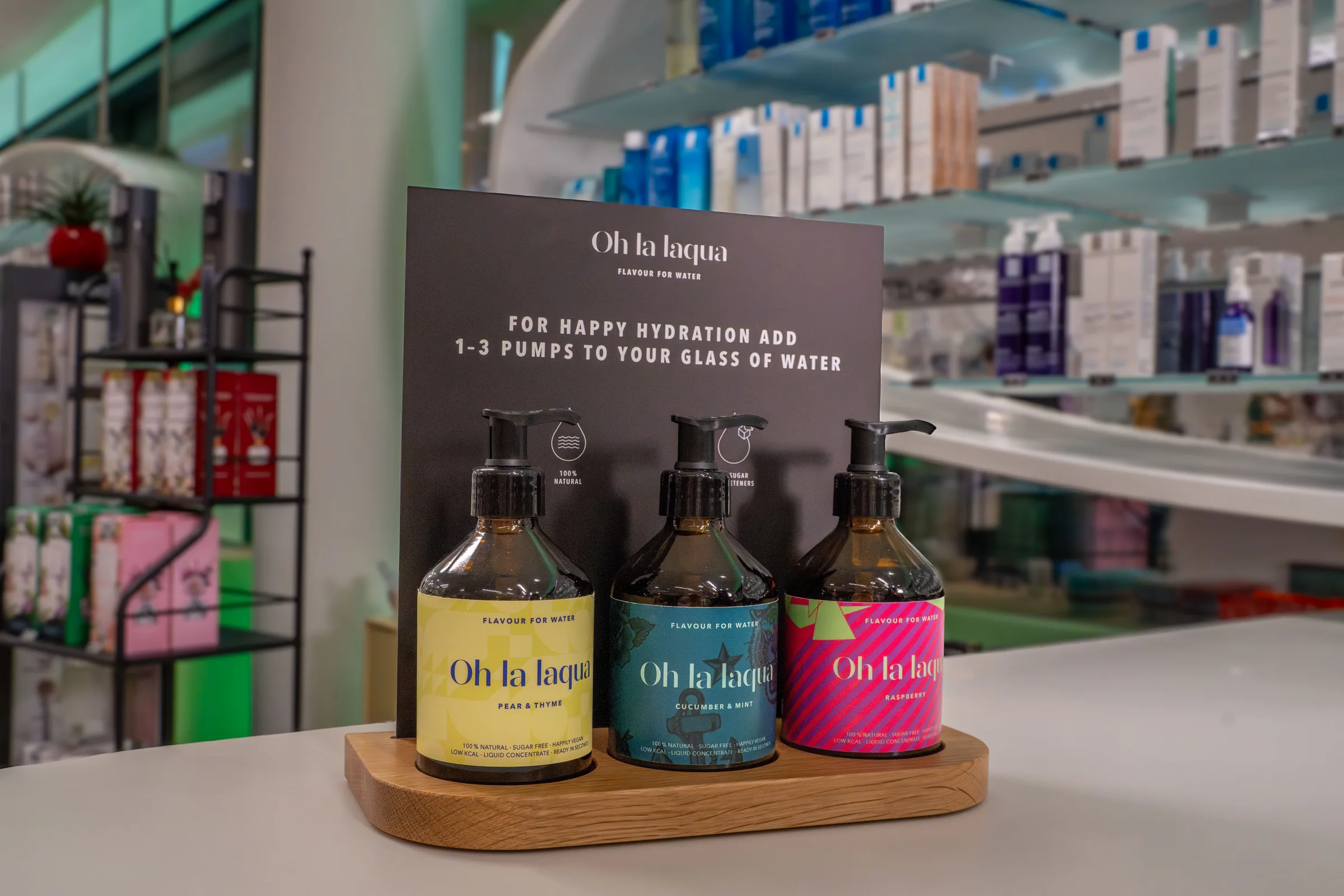 Ein Holzbrett mit drei Oh-la-laqua-Dosen in Gelb, Blau und Pink steht als Display auf der Theke einer Apotheke.