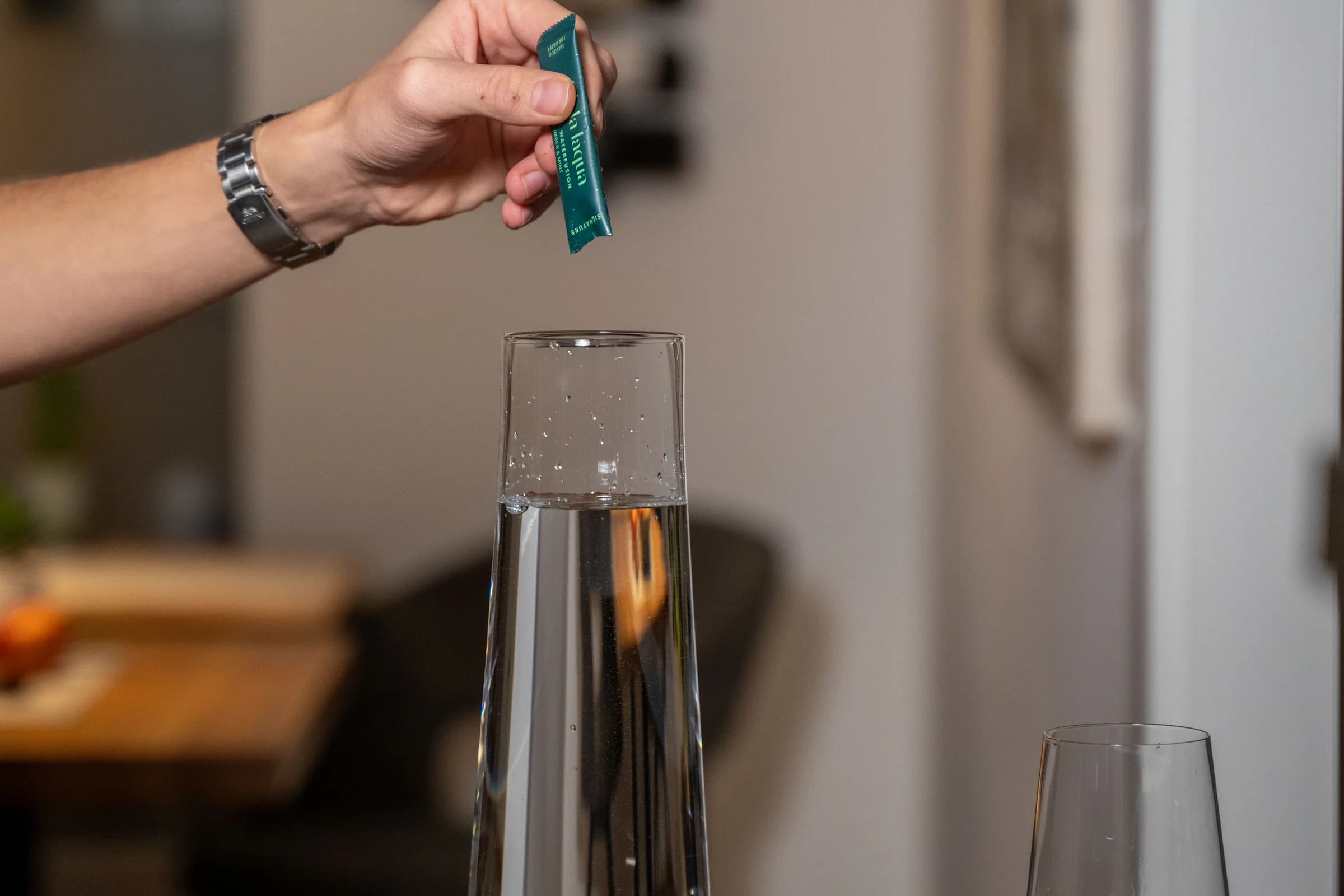 Eine Hand hält ein Oh-la-laqua-Sachet über eine Glasflasche mit Wasser, daneben ein leeres Glas.
