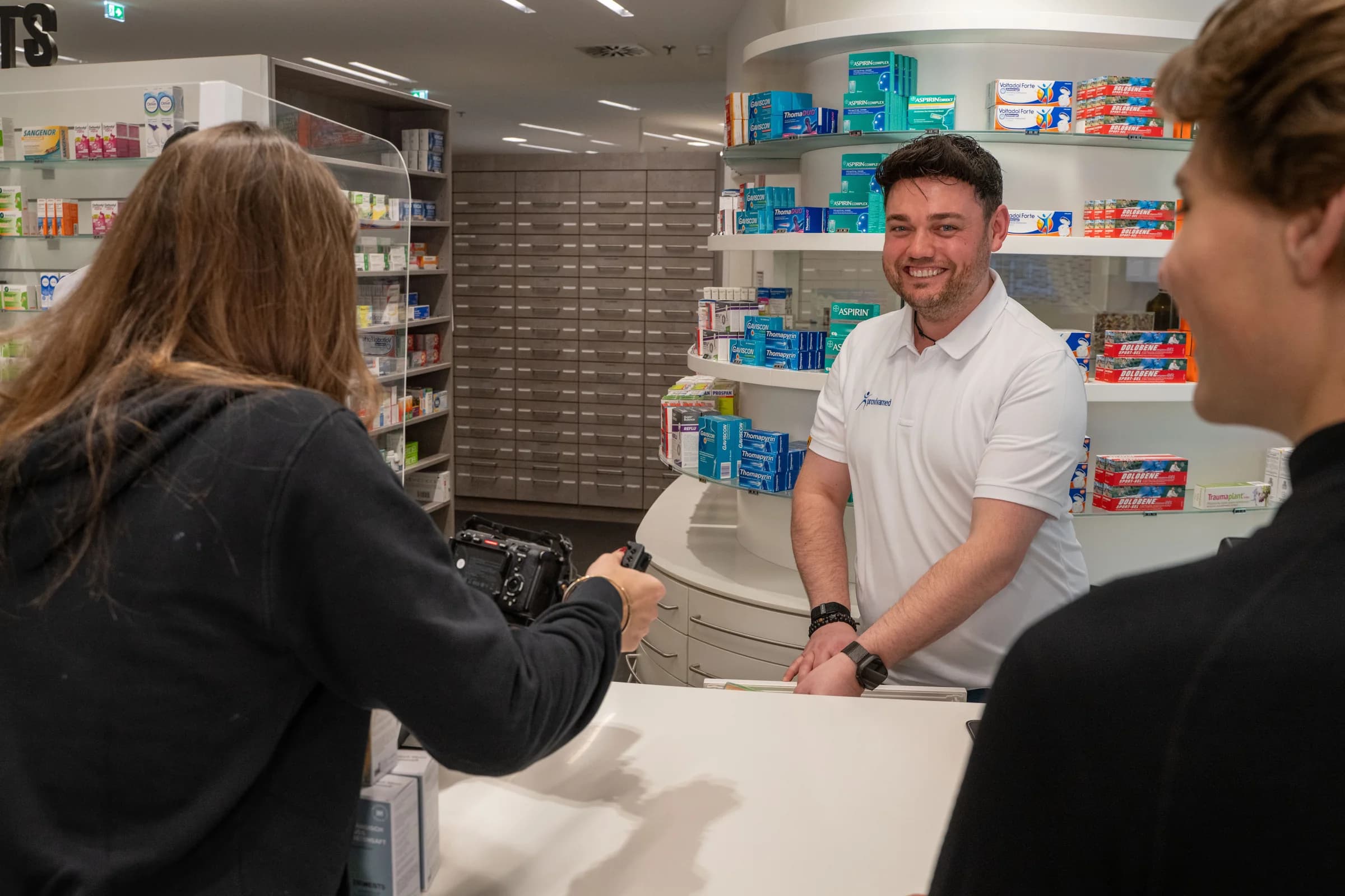 Ein Mitarbeiter im weissen provivamed-Polo bedient eine Kundin an der Theke der Apotheke, davor stehen die Produktkartons.