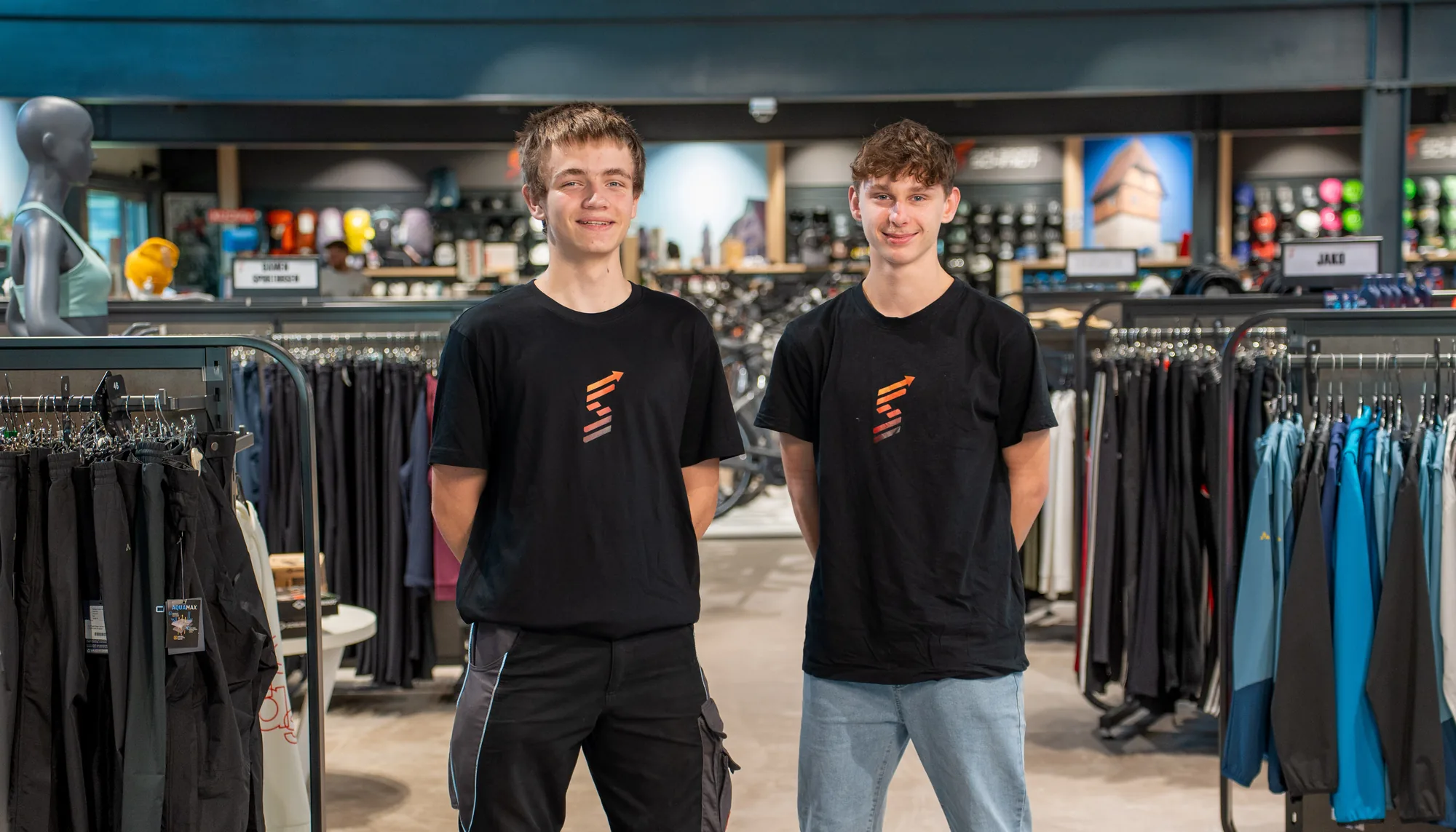 Zwei junge Mitarbeiter von Rad Sport Schmidt in schwarzen Shirts mit dem Logo des Ladens stehen lächelnd zwischen Kleiderständern, dahinter Helme und Räder.