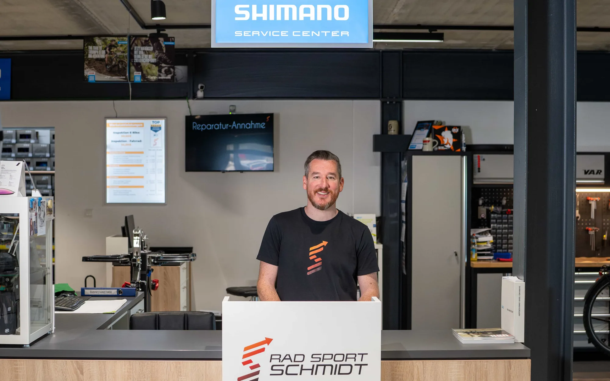Ein Mitarbeiter von Rad Sport Schmidt steht lachend an der Reparaturannahme, vor ihm die Theke mit dem Logo des Ladens, über ihm das Schild Shimano Service Center, dahinter die Werkstatt.