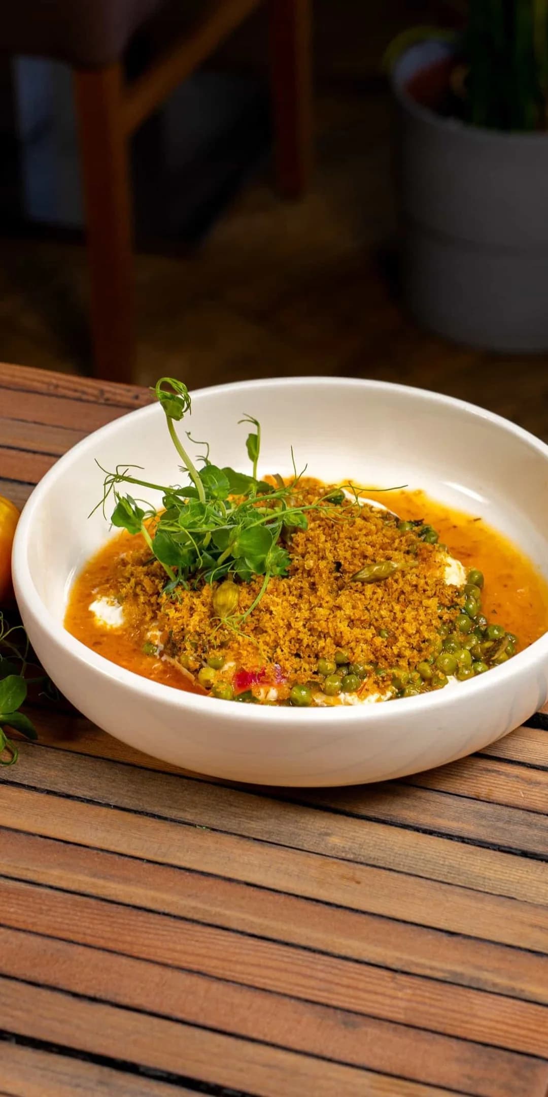 Eine weisse Schale Curry mit Erbsen, gerösteten Bröseln und Erbsensprossen neben drei gerippten Heirloom-Tomaten an der Kante eines Tischs aus Holzlatten, dahinter der Gastraum.