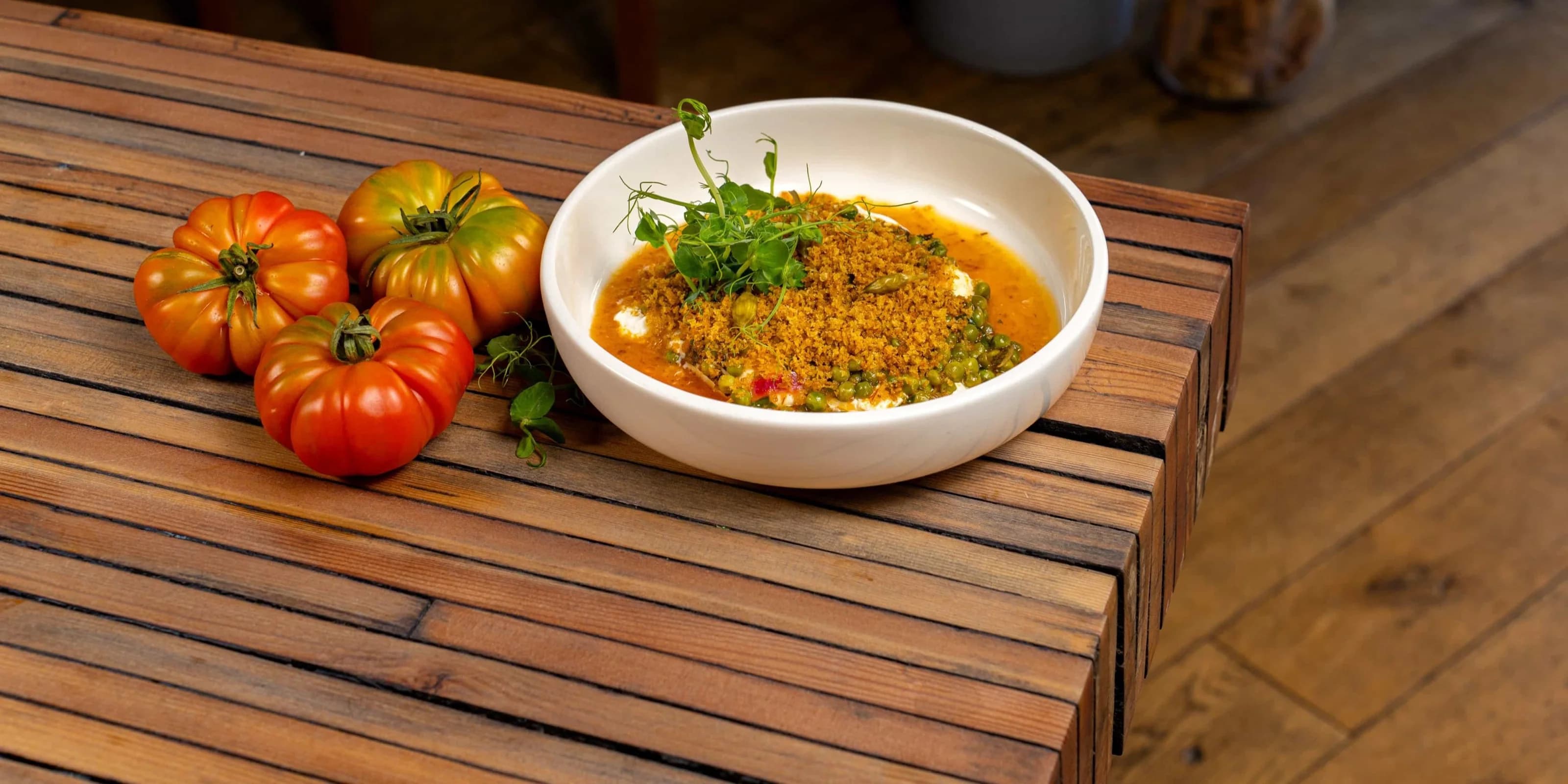 Eine weisse Schale Curry mit Erbsen, gerösteten Bröseln und Erbsensprossen neben drei gerippten Heirloom-Tomaten an der Kante eines Tischs aus Holzlatten, dahinter der Gastraum.