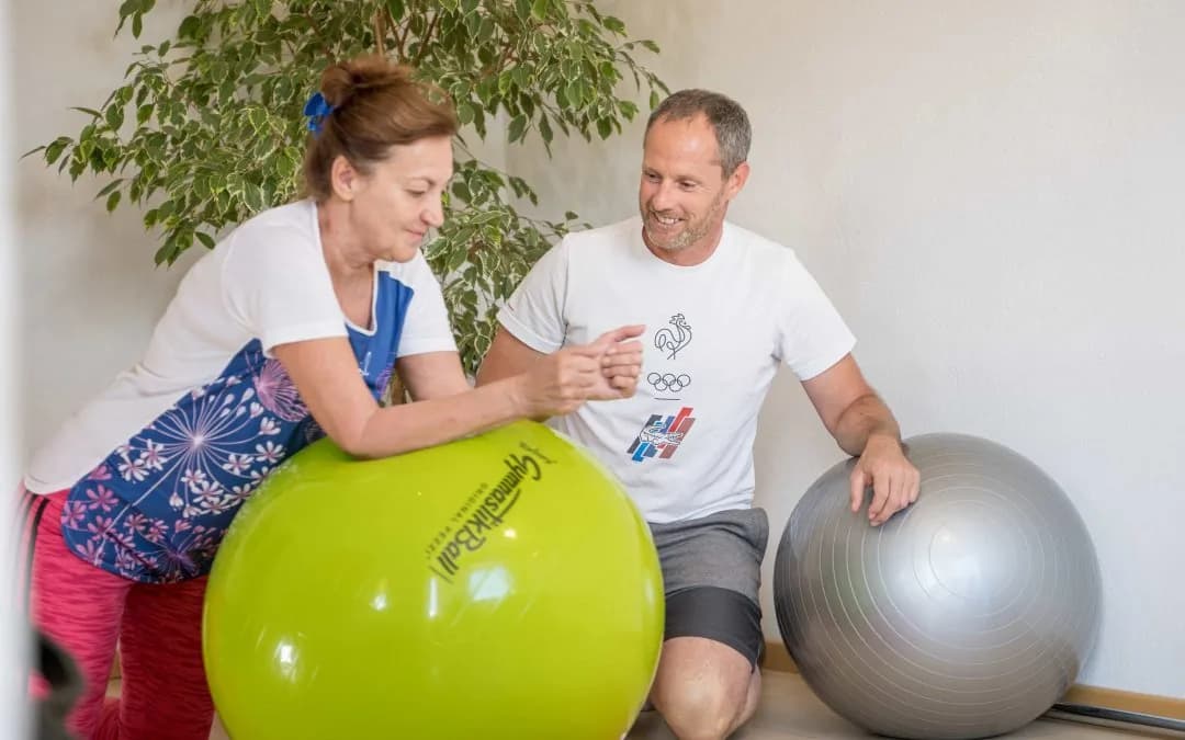 Robert Treitinger kniet neben einer Kundin, die sich auf einen grünen Gymnastikball stützt, beide lachen.