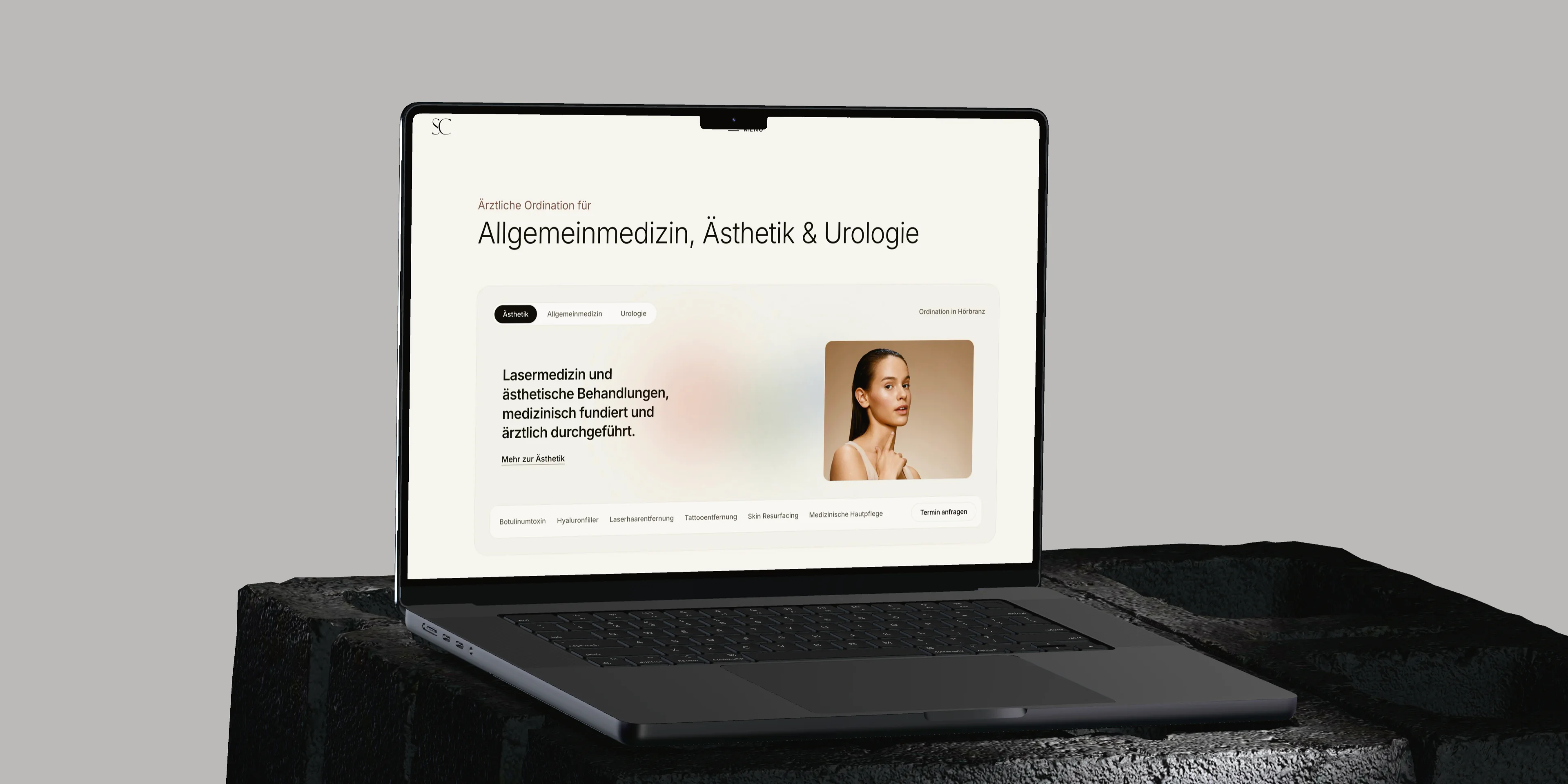 Ein MacBook auf einem dunklen Steinsockel vor hellgrauem Grund zeigt die Startseite von skinchic.at mit den drei Fachbereichen.