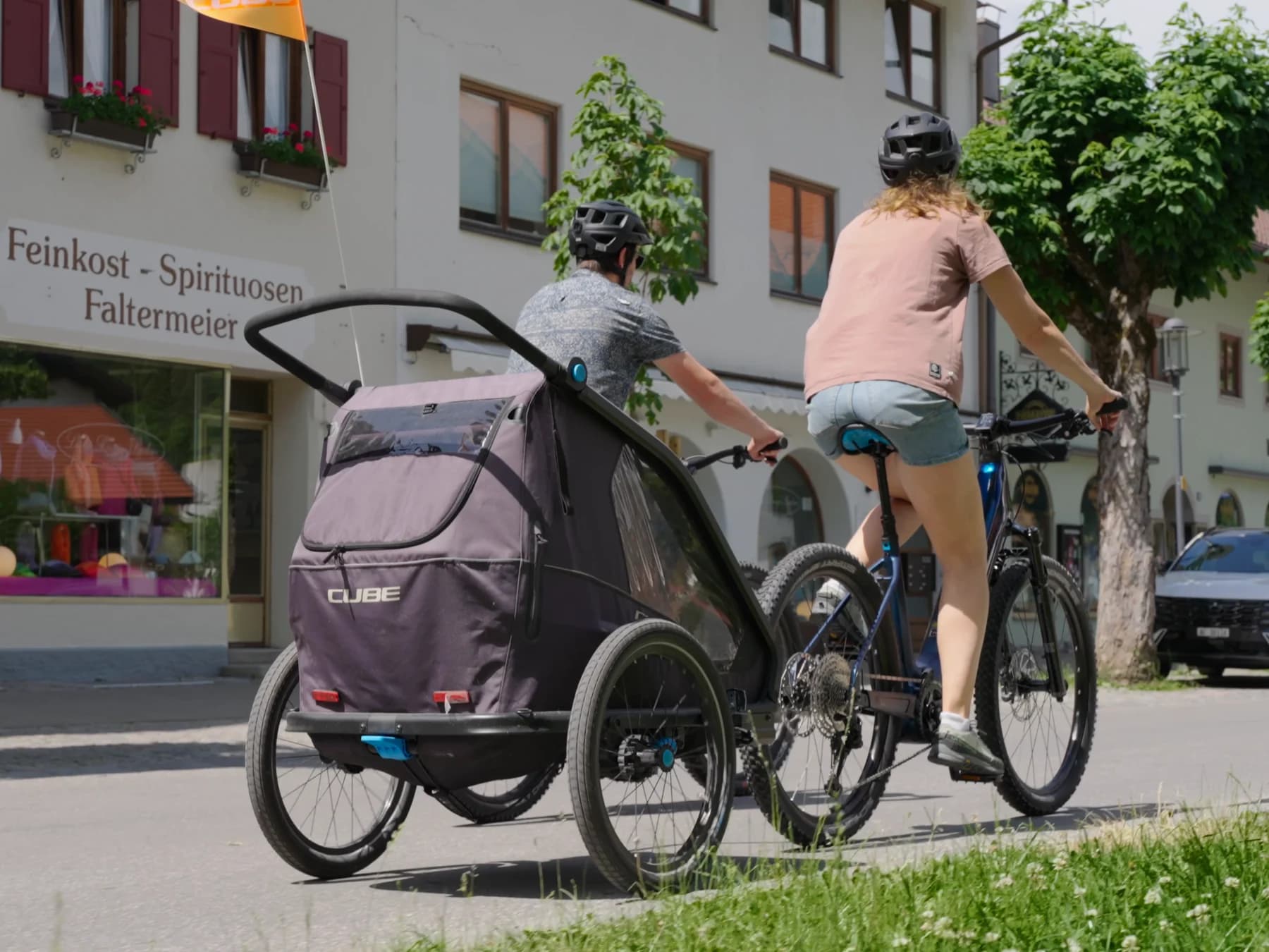 Zwei Radfahrer schieben ein Rad mit Kinderanhänger vor der Fassade des Sportgeschäfts.