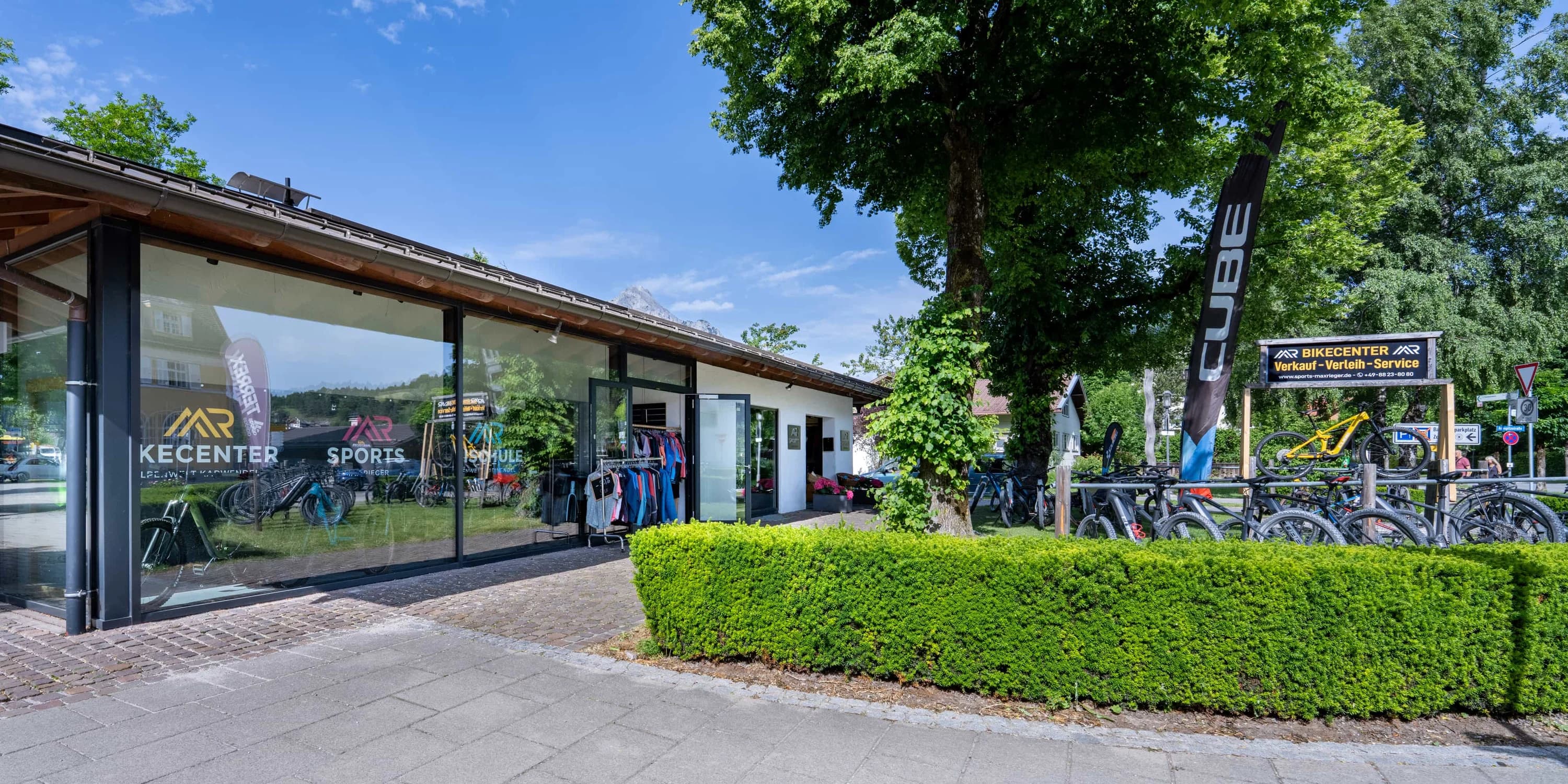 Sports Max Rieger: Die Strassenansicht des Sportgeschäfts mit Glasfront, Hecke und Baum, dahinter der Berghang.