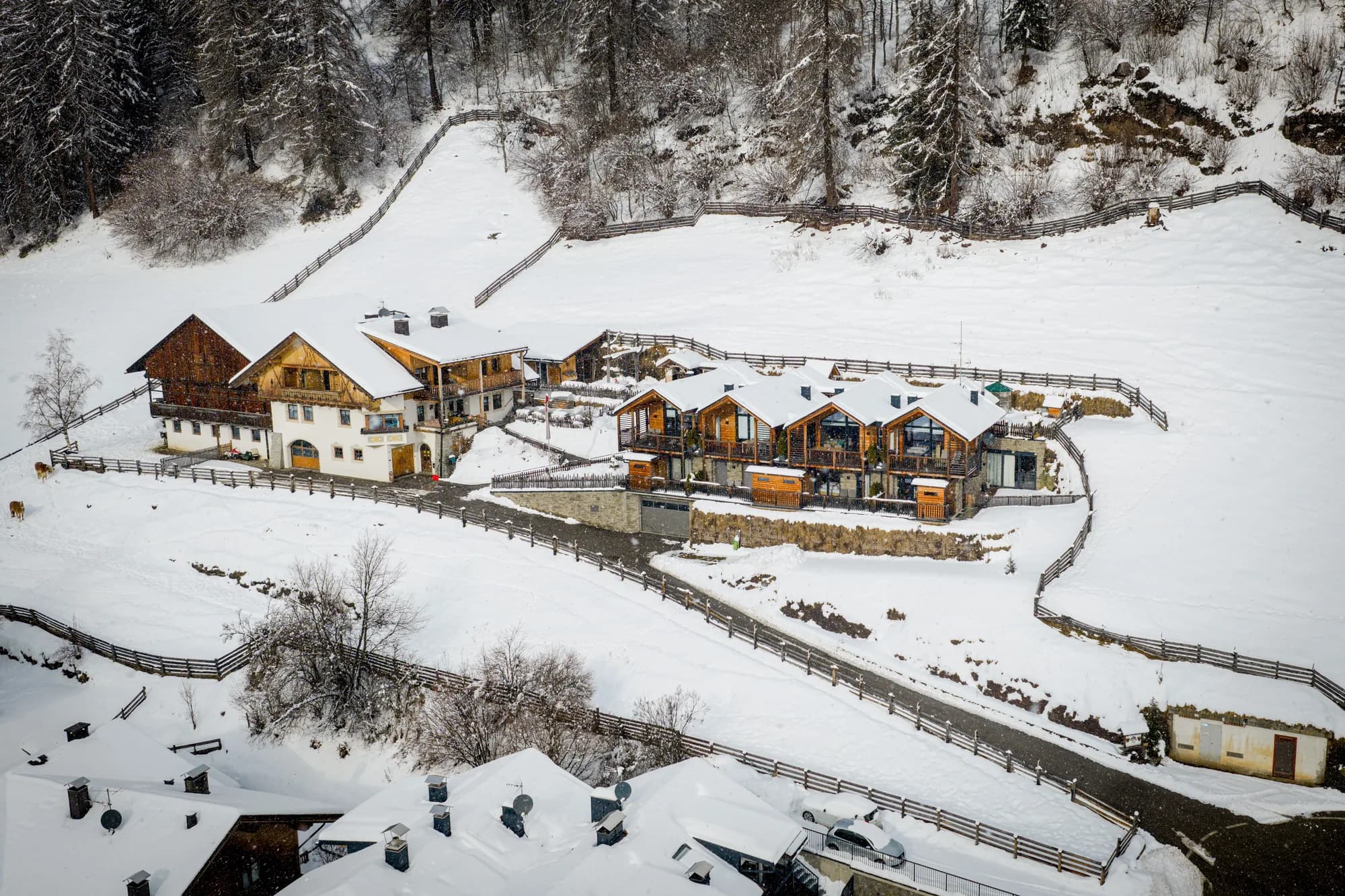 Die Zufahrt zum Stabinger Hof im Winter aus der Luft, geräumte Straße zwischen verschneiten Wiesen, rechts die Reihe der Chalets.
