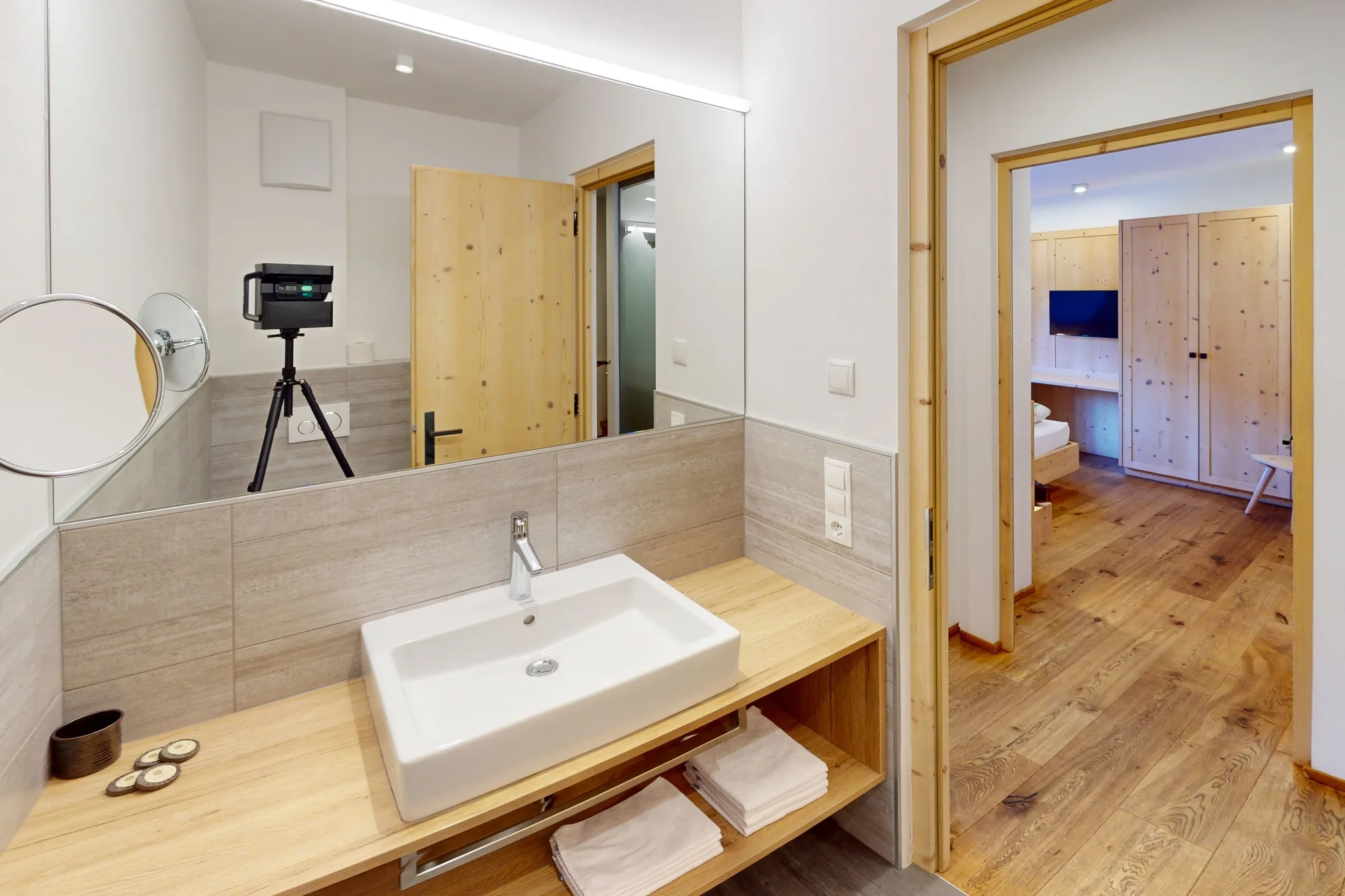Ein Bad mit Aufsatzwaschbecken auf einer Holzplatte, großem Spiegel und Kosmetikspiegel, rechts der Durchgang ins Schlafzimmer.