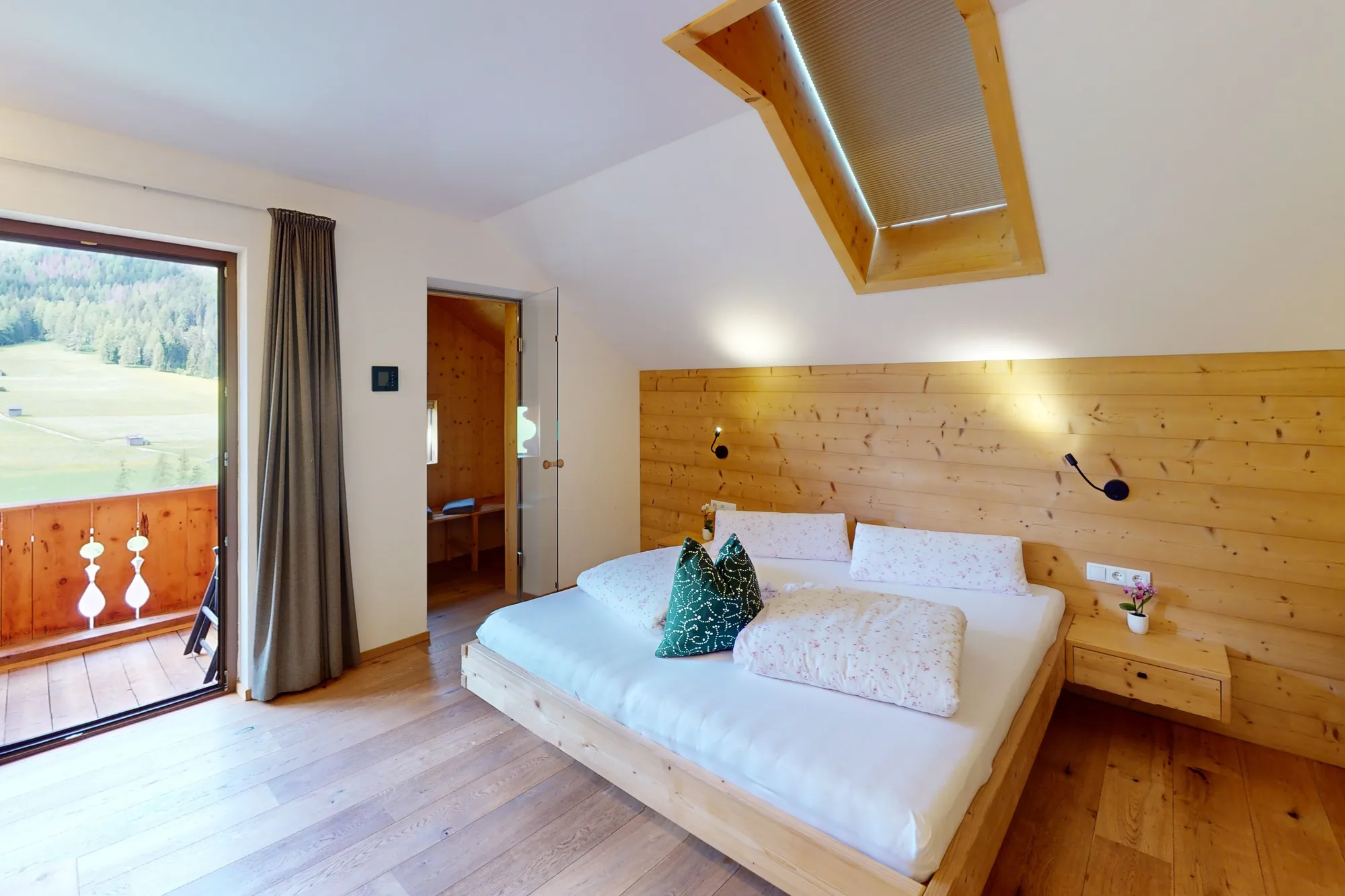 Ein Schlafzimmer mit Doppelbett vor einer Wand aus Zirbenholz, links die offene Balkontür mit Blick auf die grüne Wiese.