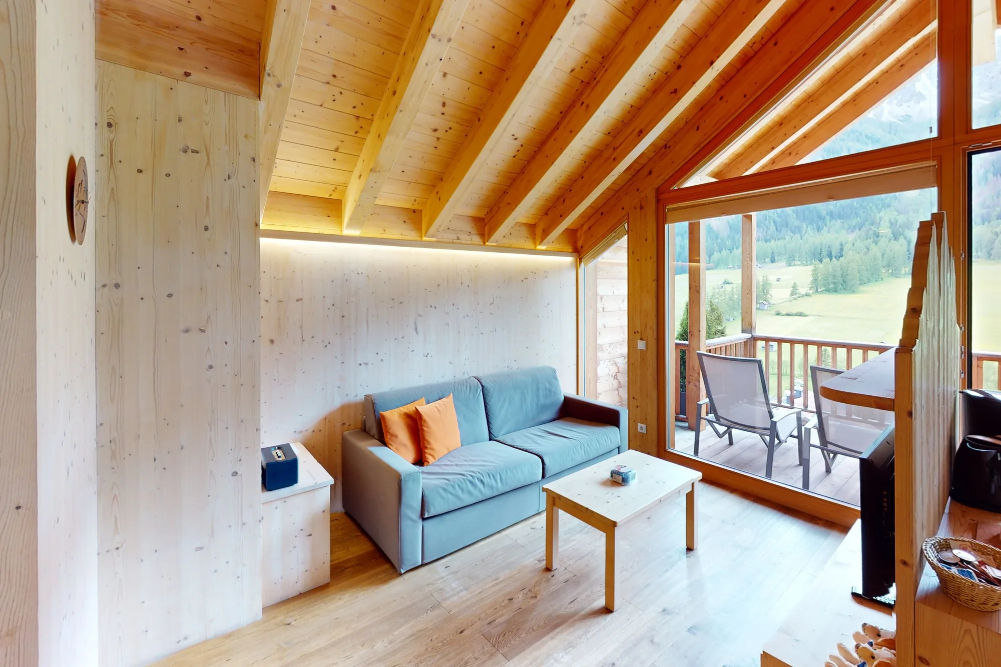 Die Stube eines Chalets unter dem hellen Holzgiebel, ein graublaues Sofa mit orangem Kissen, dahinter die Fenstertür auf den Balkon.