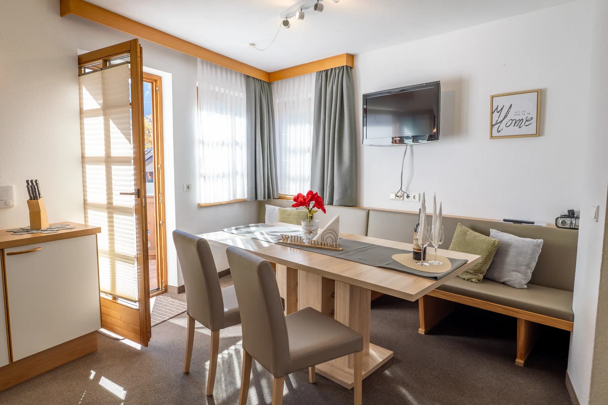 Der Essplatz der Wohnung Hias: ein heller Holztisch mit Eckbank am Fenster, rote Blumen darauf, dahinter die offene Küche.