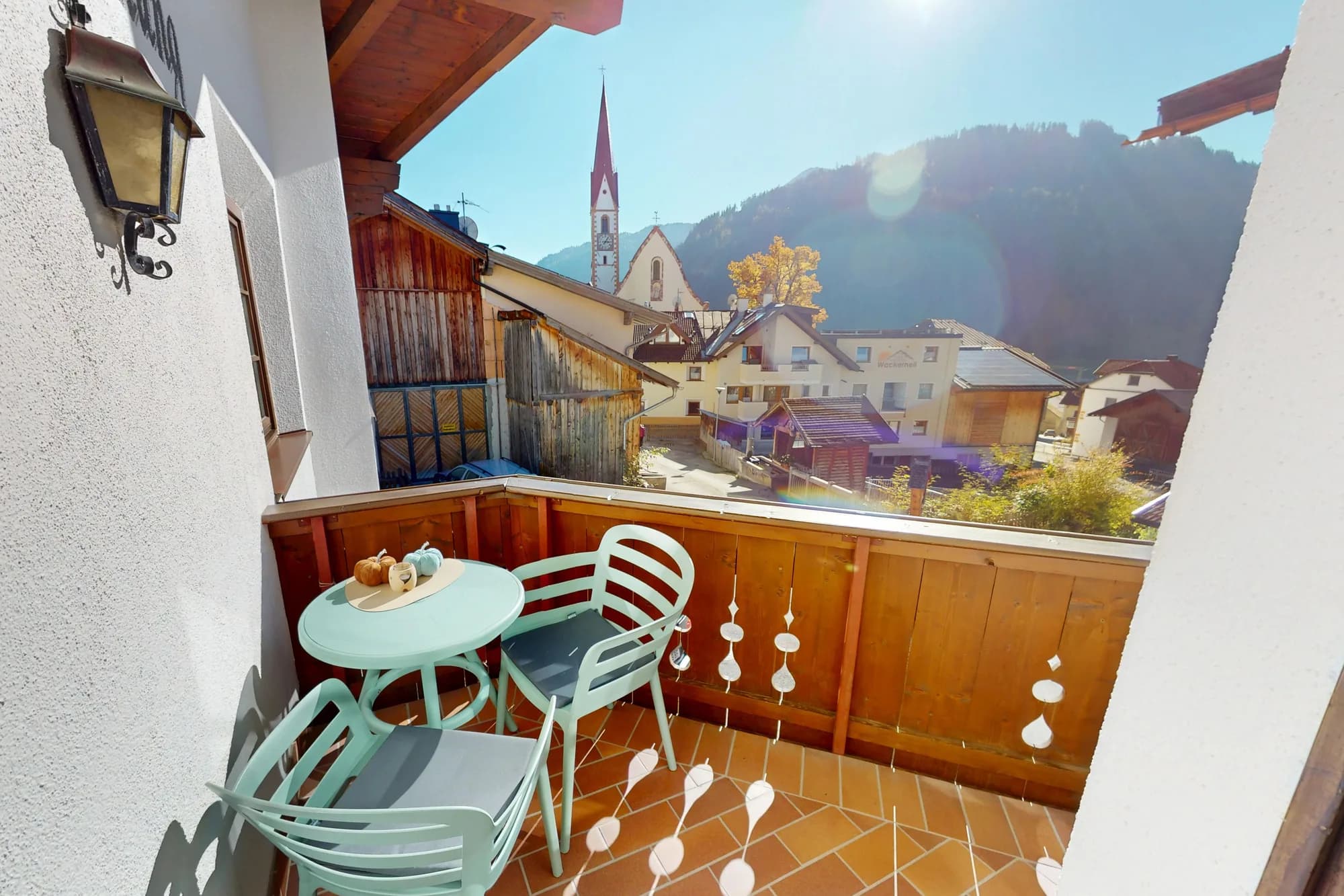Der Balkon der Wohnung Hias: ein mintgrüner Tisch mit zwei Stühlen hinter der Holzbrüstung, dahinter der Kirchturm von Nauders und die bewaldeten Hänge.