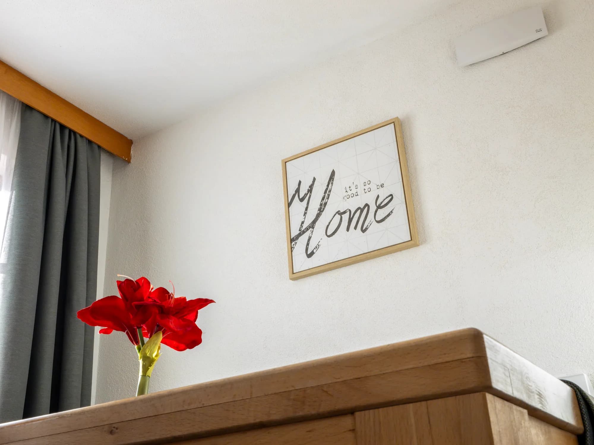 Ein gerahmter HOME-Schriftzug über dem Kopfende eines Betts, davor eine rote Amaryllis in einer schmalen Vase auf der Holzablage.