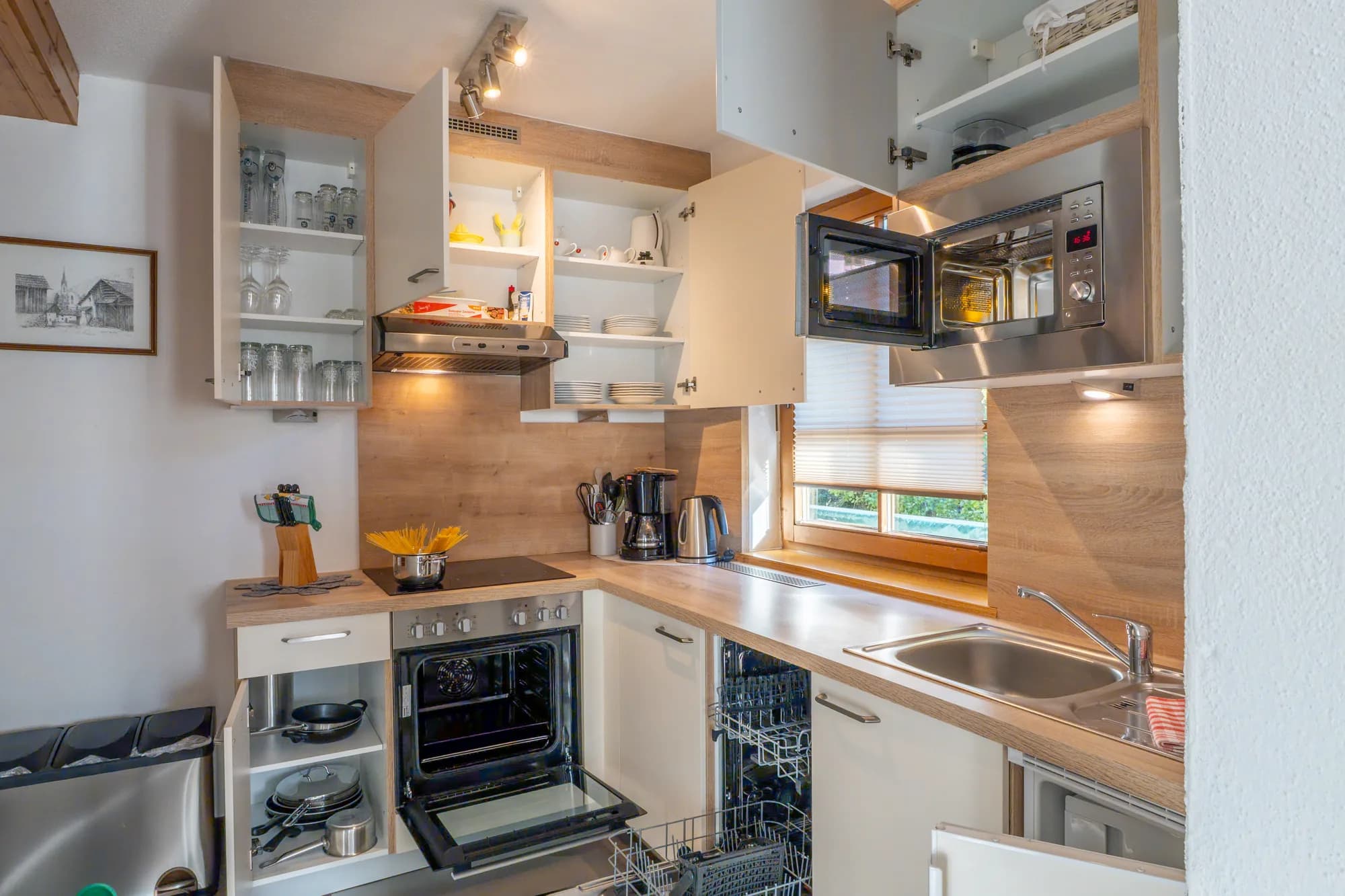 Die Küche der Wohnung BRD: weiße Fronten mit Holzarbeitsplatte, offene Hängeschränke mit Gläsern, Backofen und Spüle unter dem Fenster.