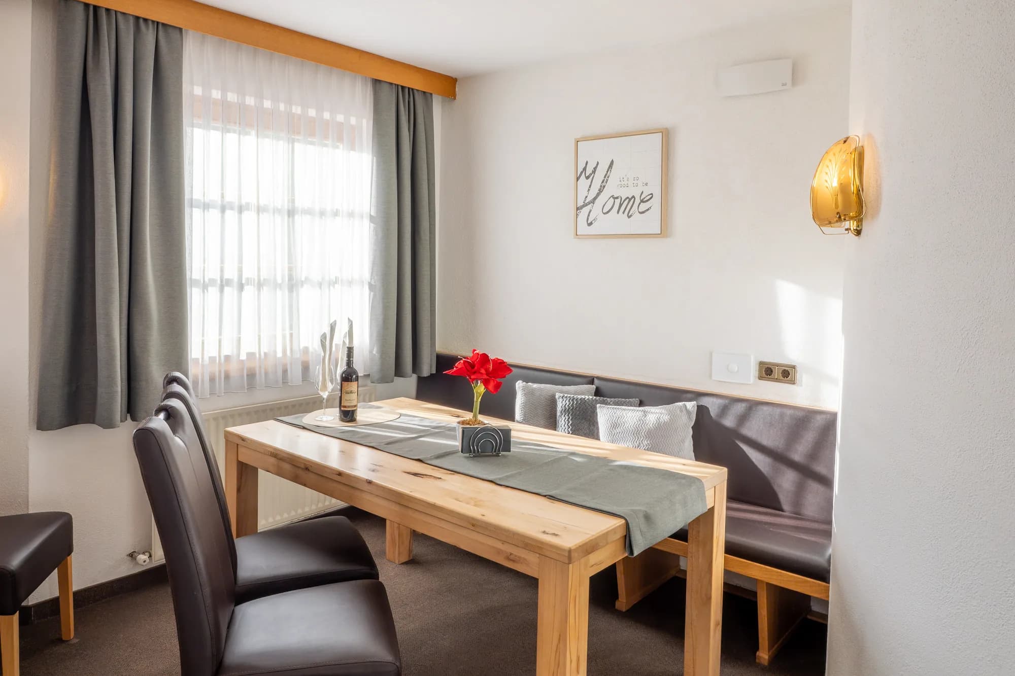 Der Essplatz der Wohnung BRD: ein langer Holztisch mit Eckbank und Stühlen vor dem Fenster, an der Wand ein gerahmter HOME-Schriftzug, daneben rote Blumen.