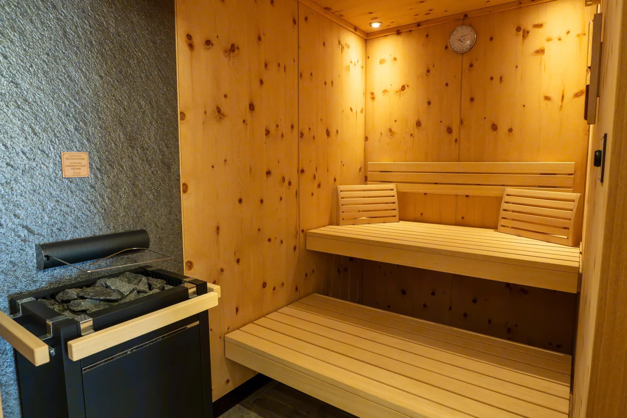Die Zirbensauna der Wohnung Hias: helle Holzbänke über Eck, links der schwarze Ofen mit Steinen, an der Wand eine kleine Sanduhr.