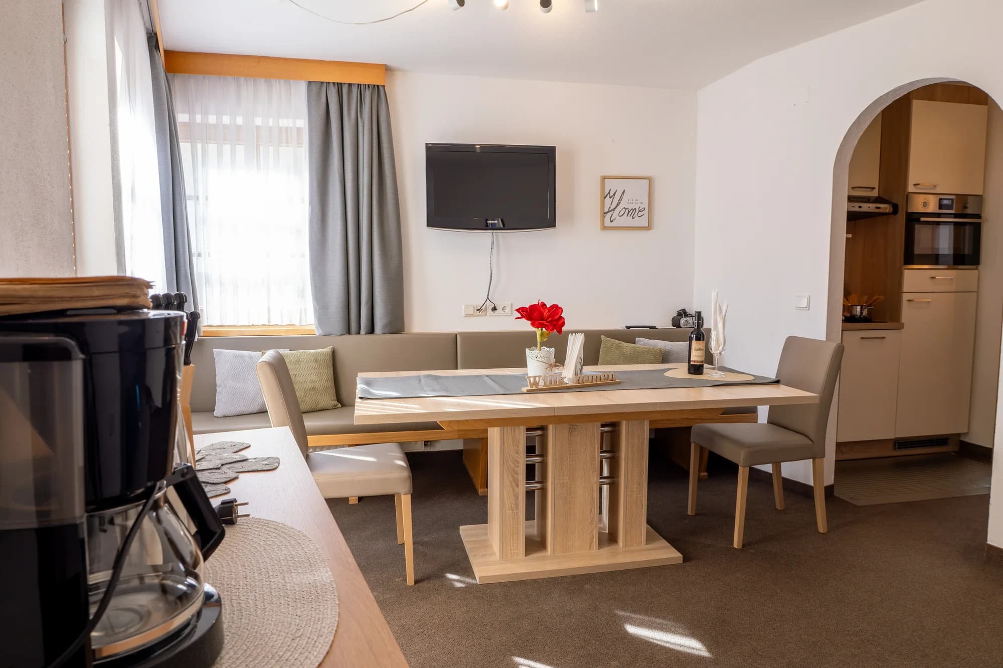 Die Stube der Wohnung Hias: ein schwarzer Kaminofen im Vordergrund, dahinter Esstisch mit Bank, Fernseher an der Wand und der Durchgang zur Küche.