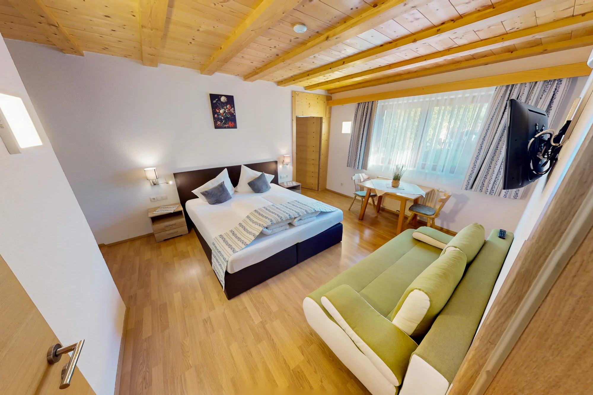 Der offene Wohnraum der Wohnung BRD unter dem hellen Holzgiebel: ein Doppelbett links, daneben ein grünes Sofa und ein Fenster mit Blick auf den Hang.