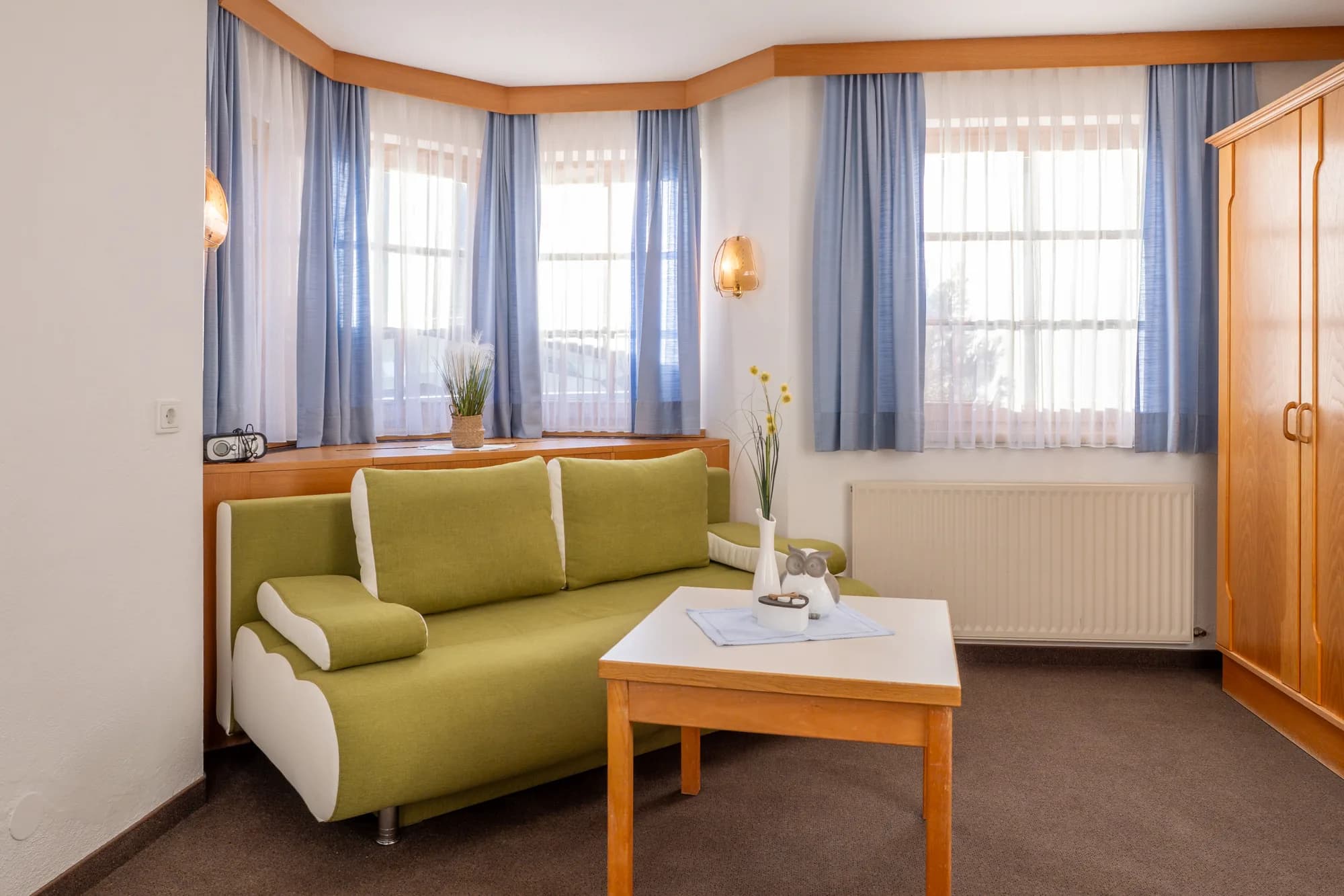 Der Wohnraum der Wohnung Stadl: ein grünes Sofa mit Holztisch vor drei Fenstern mit blauen Vorhängen, rechts ein Kleiderschrank aus Naturholz.