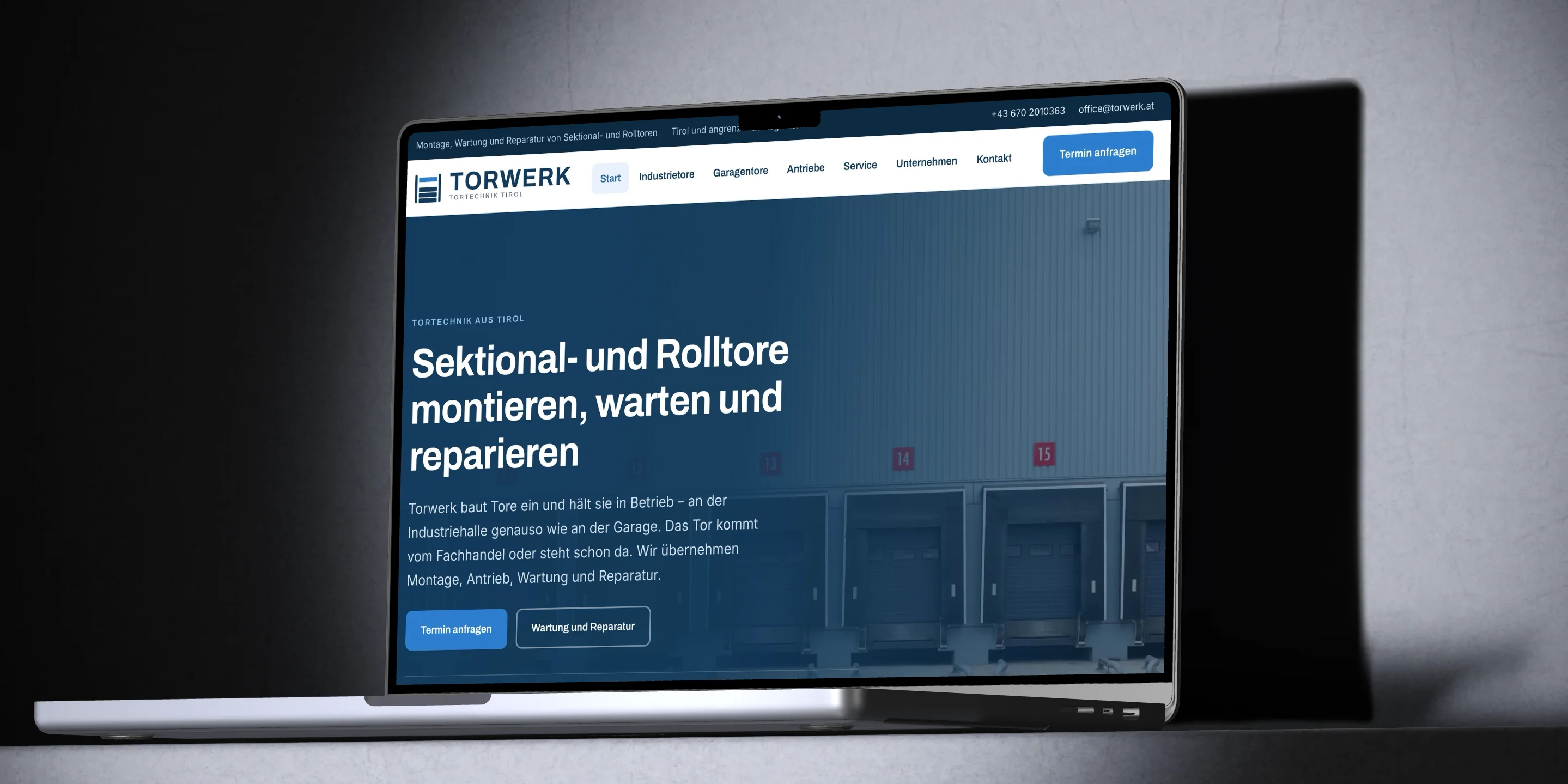 Ein aufgeklapptes MacBook auf einer grauen Kante im Lichtkegel zeigt die Startseite von torwerk.at mit der Überschrift Sektional- und Rolltore montieren, warten und reparieren.
