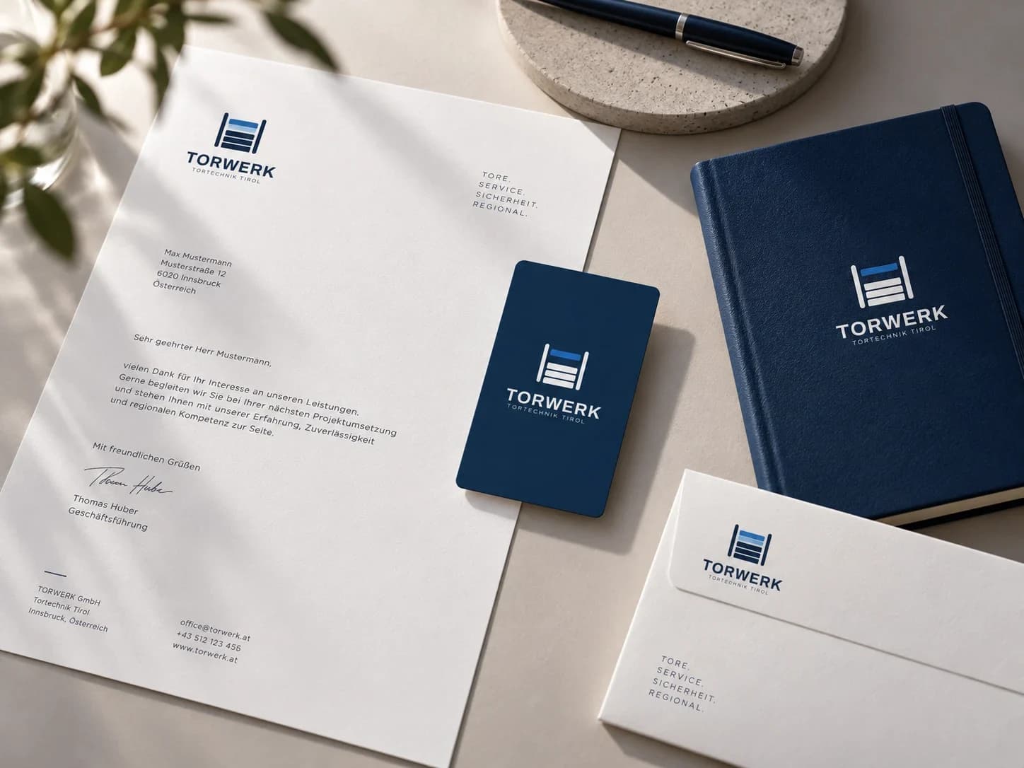 Geschäftsausstattung von Torwerk Tirol auf einem hellen Tisch: ein Briefbogen mit Logo, eine dunkelblaue Visitenkarte, ein blaues Notizbuch mit dem Signet und ein weißes Kuvert, daneben ein Füller.