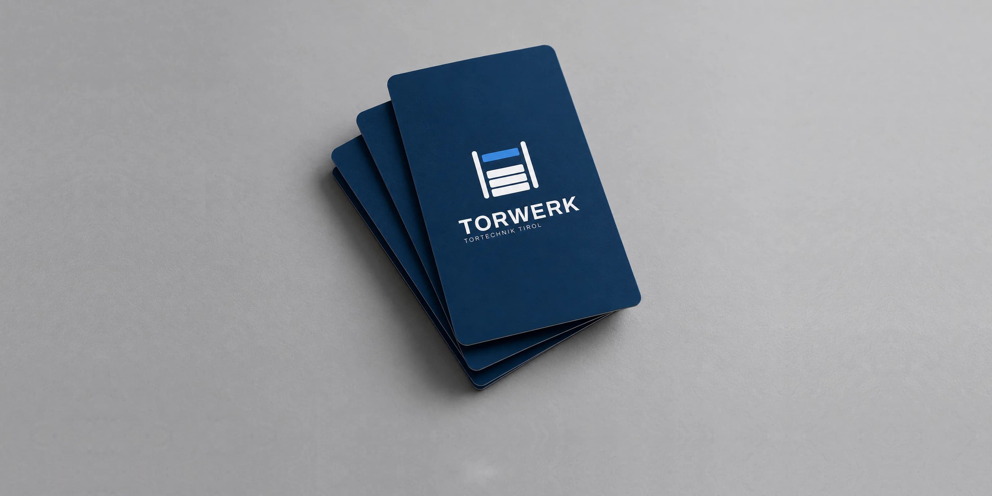 Aufgefächerte Visitenkarten von Torwerk Tirol im dunklen Markenblau auf grauem Grund: das Signet aus zwei Schienen und vier Lamellen, darunter der Schriftzug Torwerk und die Zeile Tortechnik Tirol.