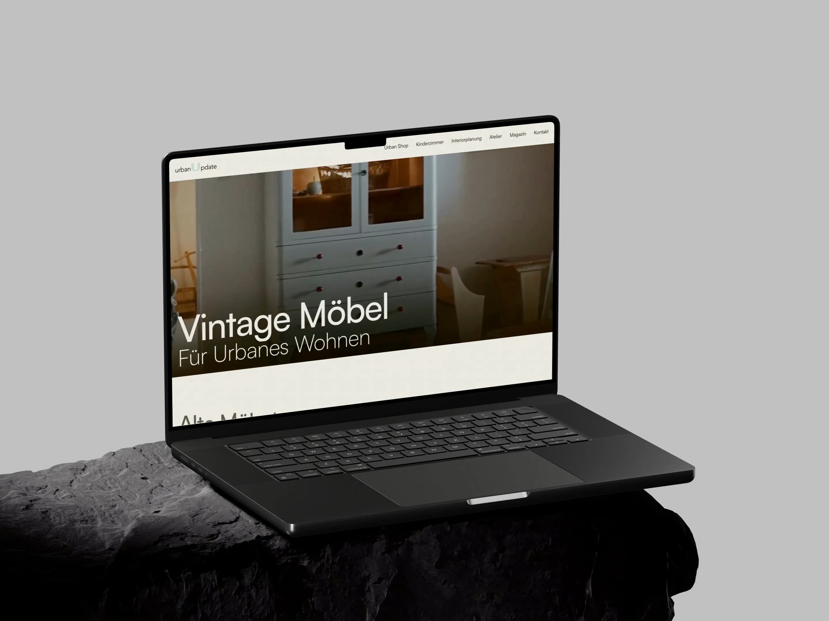 UrbanUpdate: Ein aufgeklapptes MacBook Pro auf einem dunklen Steinsockel zeigt die Startseite von urbanupdate.at mit einem hellblauen Vintage-Kasten und der Zeile Vintage Möbel für urbanes Wohnen.