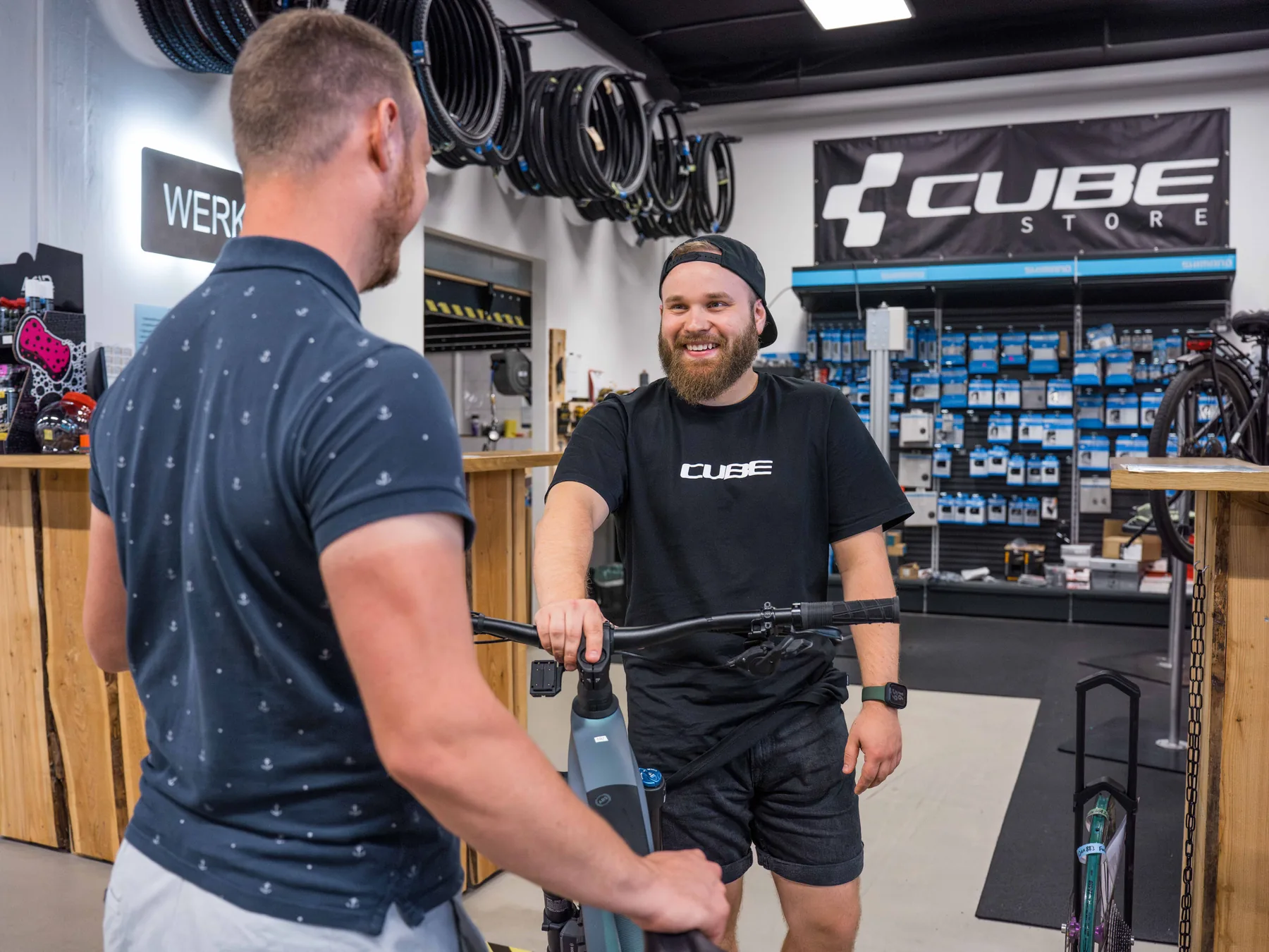 Übergabe vor der Werkstatt im CUBE Store Hildesheim: Ein Mitarbeiter im schwarzen CUBE-Shirt hält ein Rad und lacht einen Kunden an, dahinter das CUBE-Store-Banner und die Wand mit Ersatzteilen.