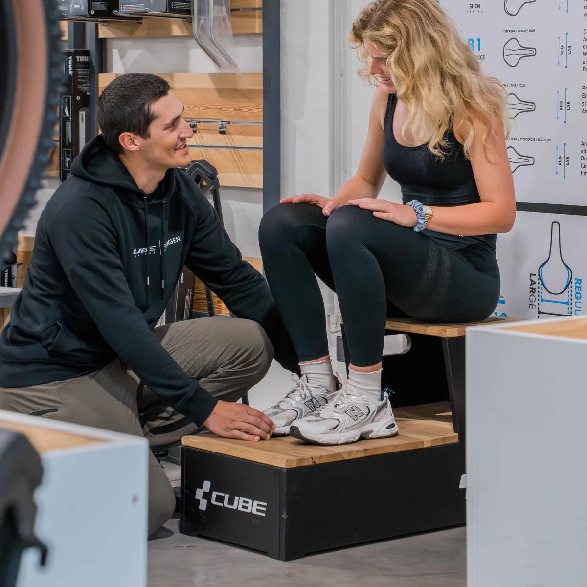 Sitzknochenvermessung im CUBE Store Göttingen: Ein Mitarbeiter kniet vor einer Kundin, die auf einer Messbank sitzt, daneben eine Tafel mit Sattelgrößen.