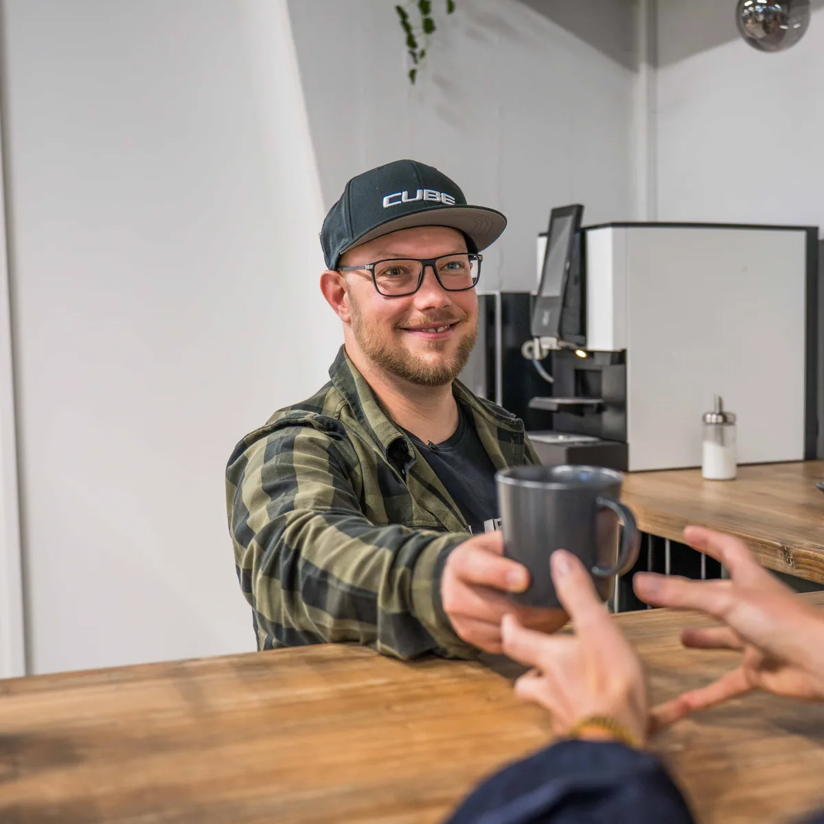Ein Mitarbeiter mit Brille und CUBE-Kappe reicht einer Kundin am Holztresen des CUBE Store Hildesheim eine Tasse Kaffee.