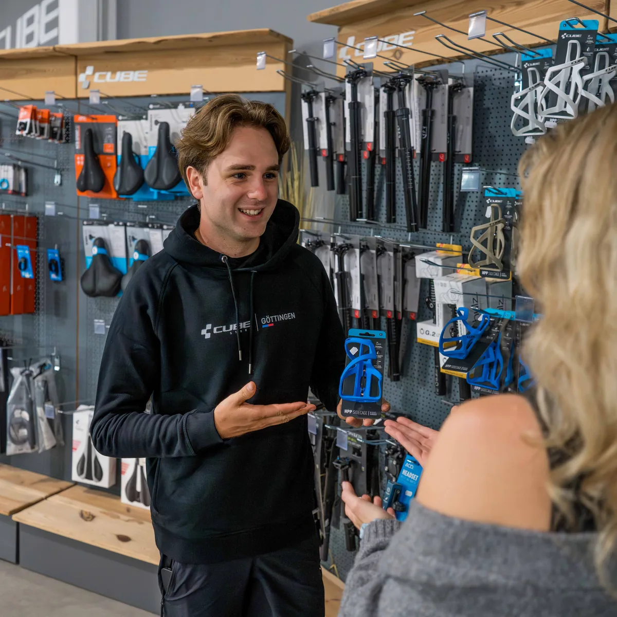 Ein Mitarbeiter im Store-Hoodie berät eine Kundin an der Zubehörwand des CUBE Store Göttingen und zeigt ihr einen Flaschenhalter.