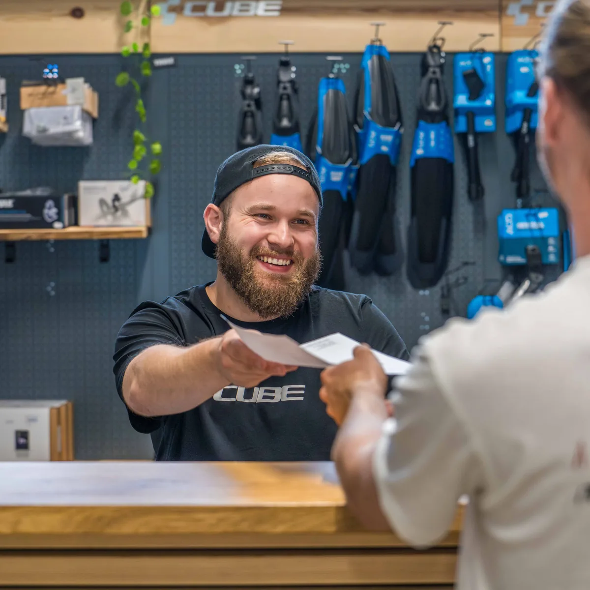 Ein Mitarbeiter im CUBE-Shirt reicht einem Kunden an der Kasse des CUBE Store Hildesheim lachend einen Beleg über den Tresen.